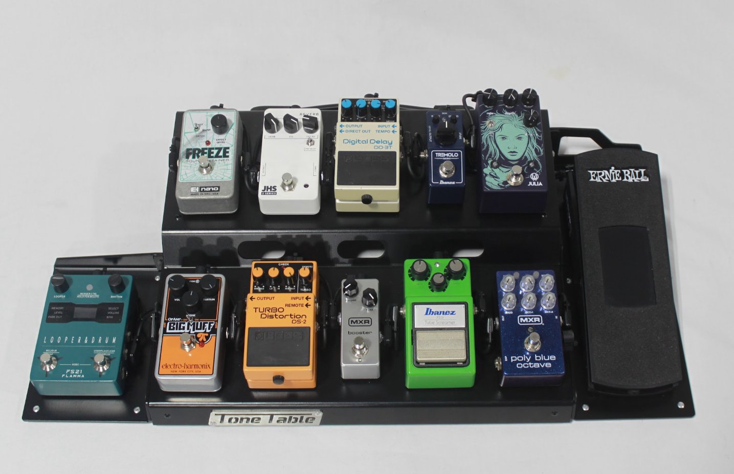 A guitar effects pedalboard with various guitar effect pedals and a power supply mounted on a black Tone Table pedalboard with a foot pedal. Pedals include reverb, delay, overdrive, distortion, tuner, looper, and modulation effects in different color