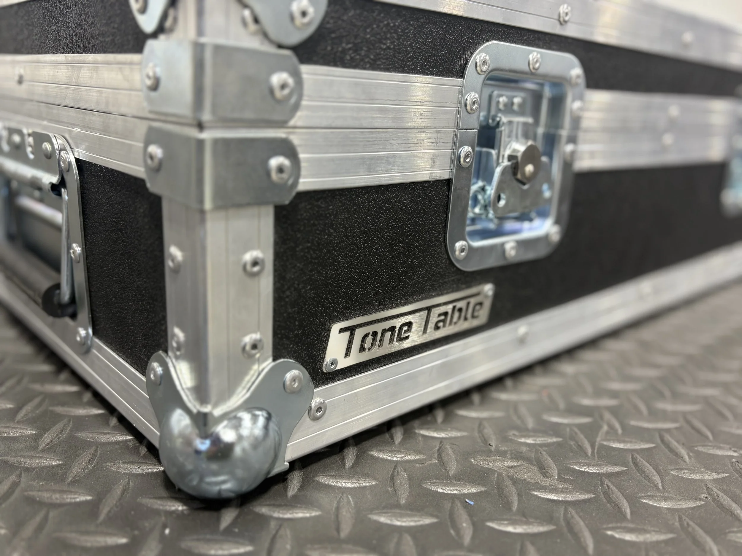 Close-up of a black, metal flight case with metal reinforcements, a latch, and a TONE TALLIE logo on the front, placed on a textured metal surface.