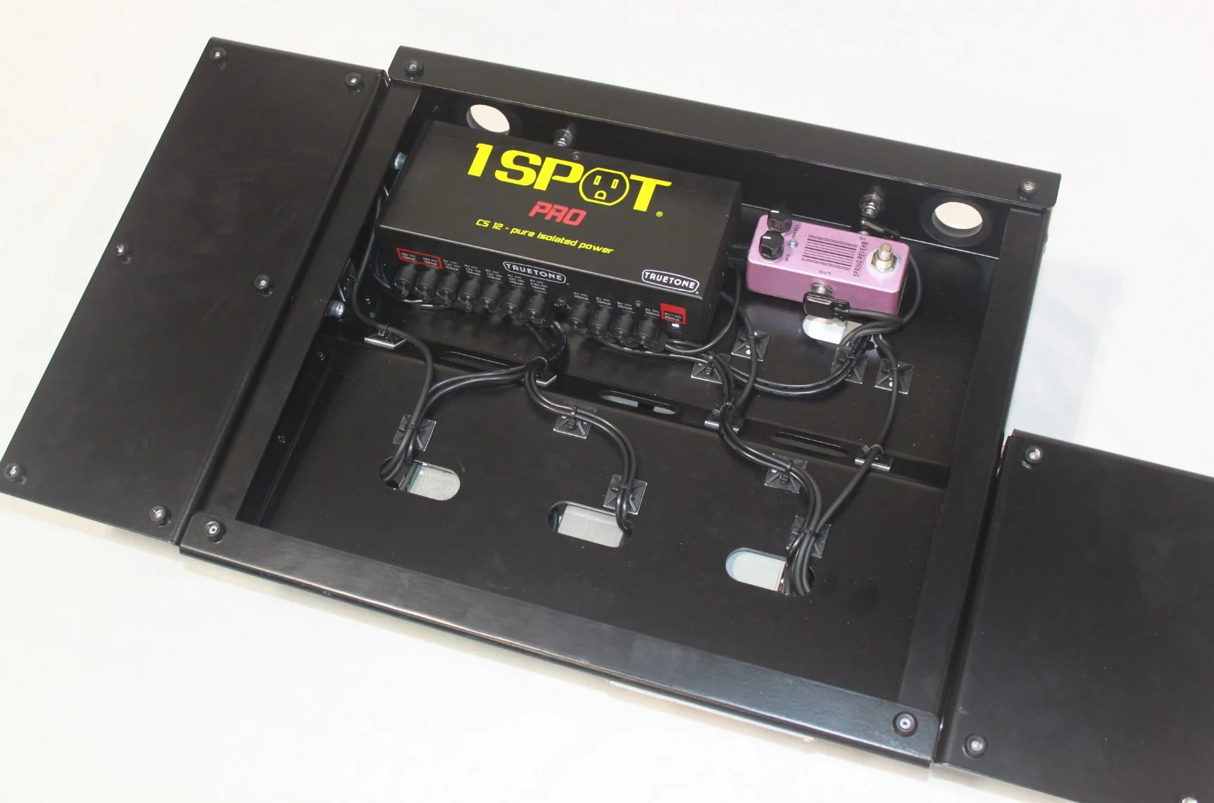 Interior of a black pedalboard with various electronic components, including a black 1Spot power supply, a purple effects pedal, some cables, and connectors.