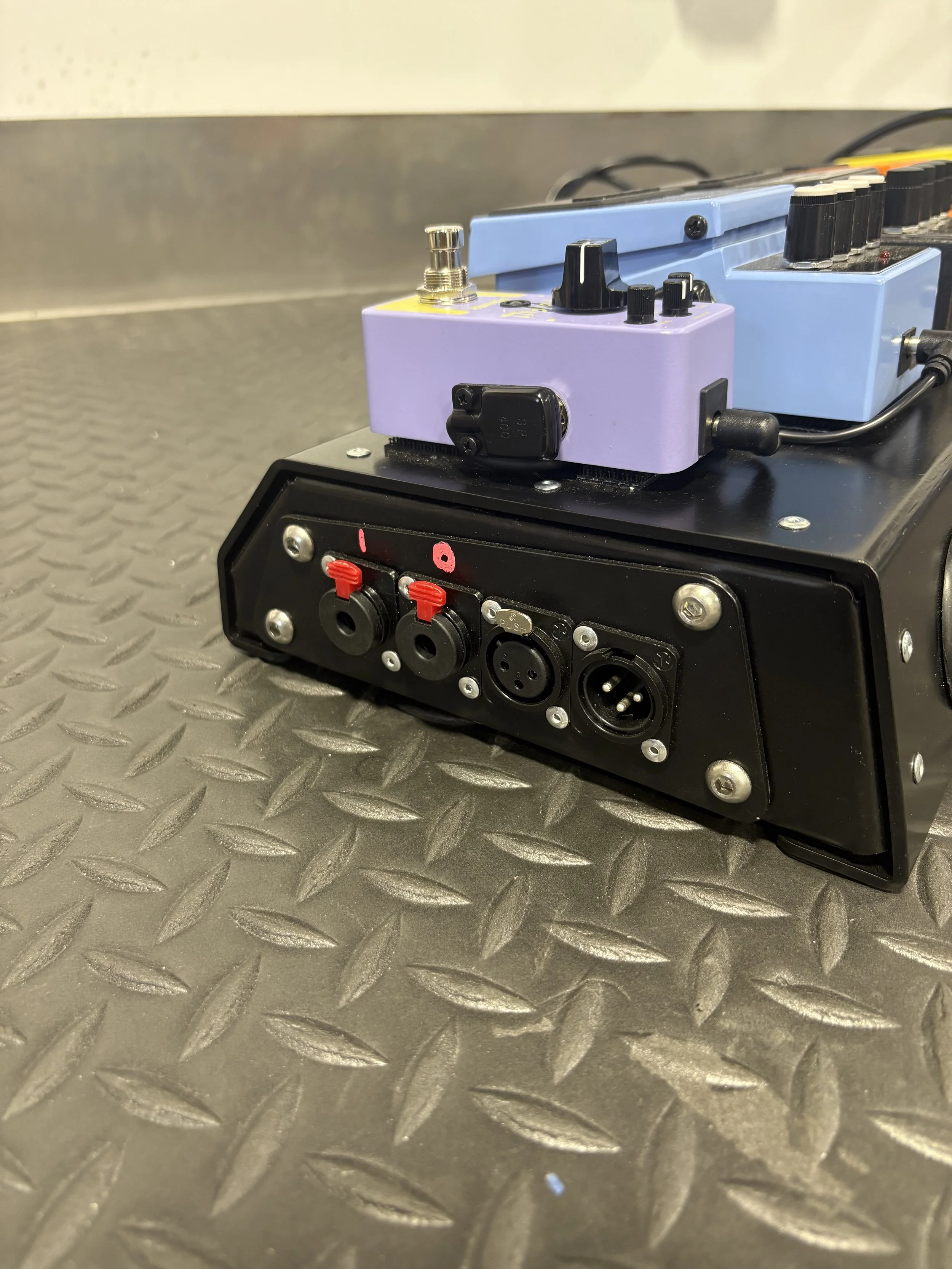 Close-up of a black pedalboard with various guitar effect pedals, including purple and blue pedals, on a metallic textured surface.