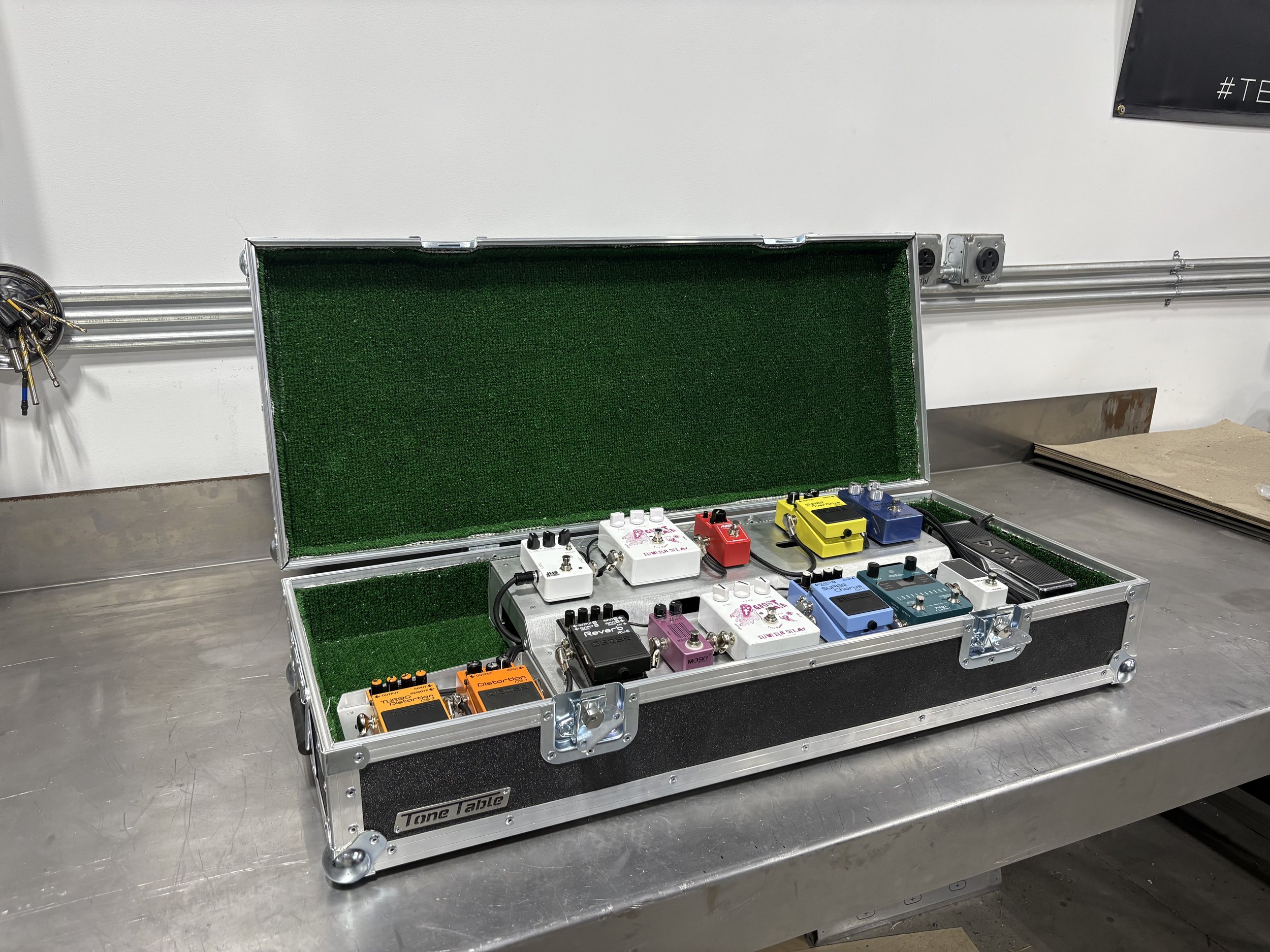 Open black and silver guitar effects pedal case with green interior containing multiple guitar pedals of various colors, including orange, purple, blue, yellow, white, and black, on a metal workbench.