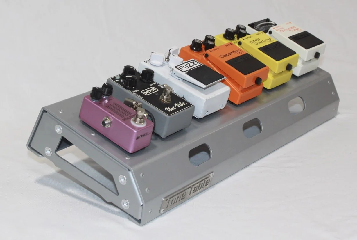 Six guitar effect pedals in pink, black, white, orange, yellow, and white colors on a metal pedalboard.