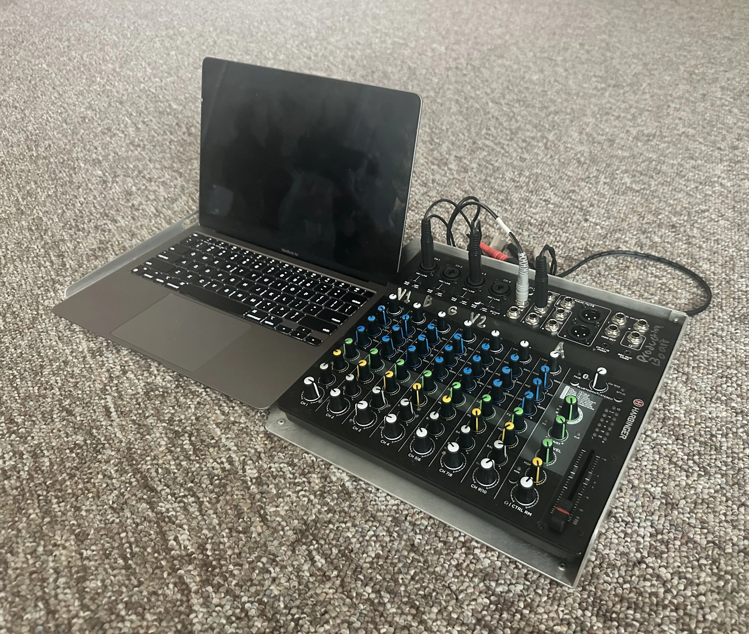 A digital audio mixer with colorful knobs and sliders, connected to a MacBook laptop, all placed on a carpeted floor.