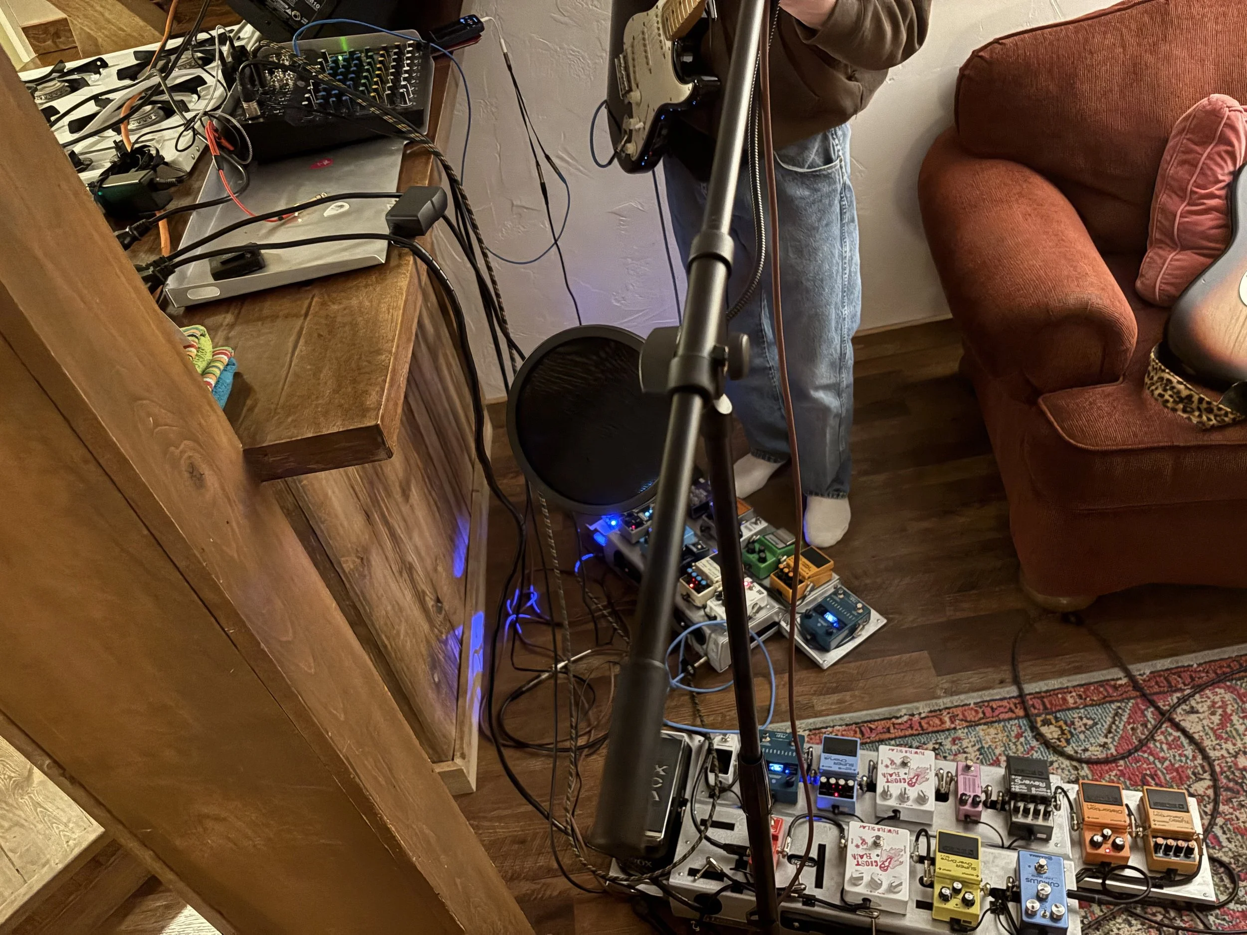 Close-up of an electric guitar on a stand with guitar effect pedals on the floor, a microphone stand, and a person standing nearby with part of their body visible. A wooden side table with electronic equipment and cables is also in the scene, with a 