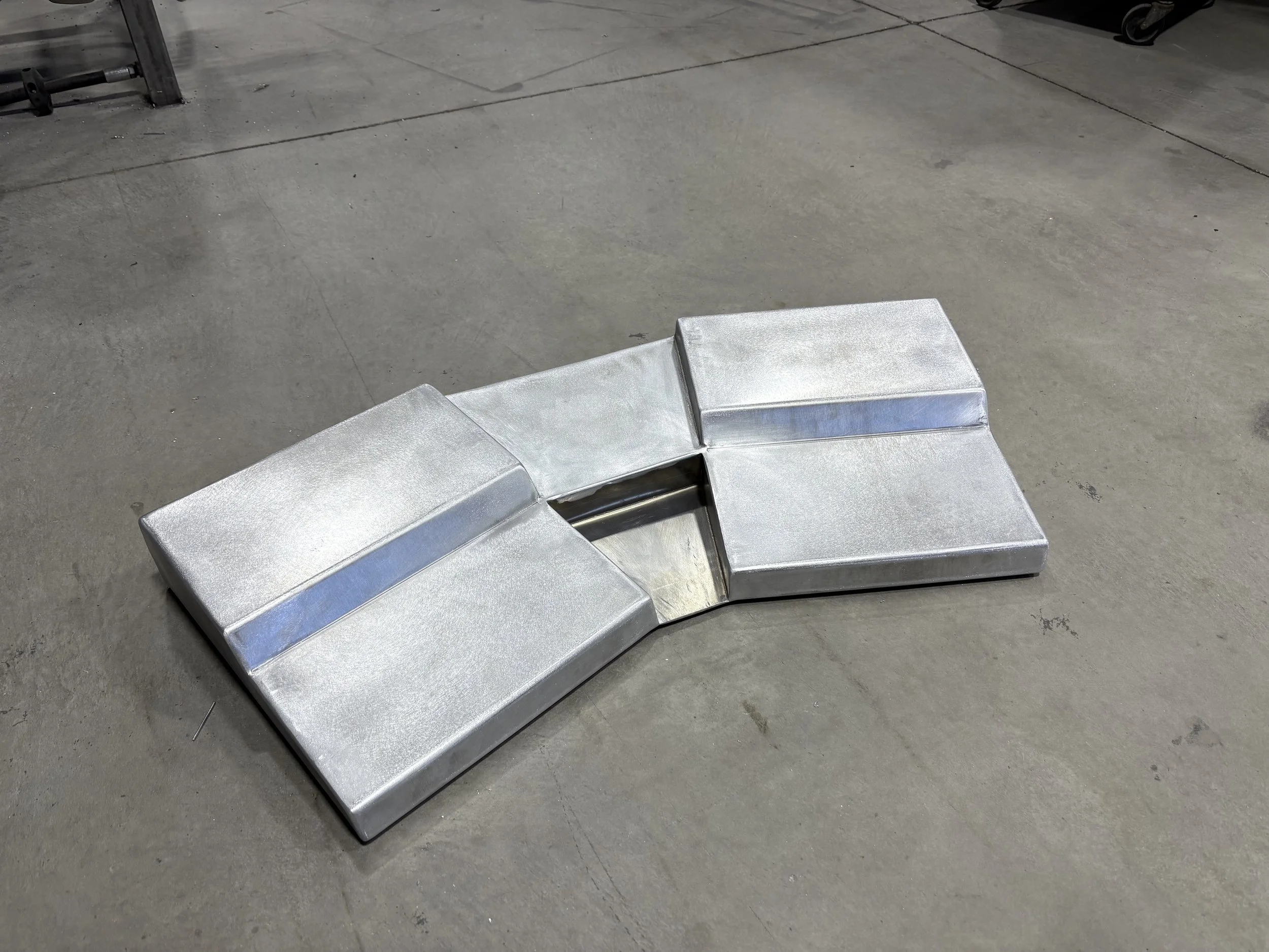 A metallic, modern-style bench with four separate sections connected by a central opening, placed on a concrete floor.