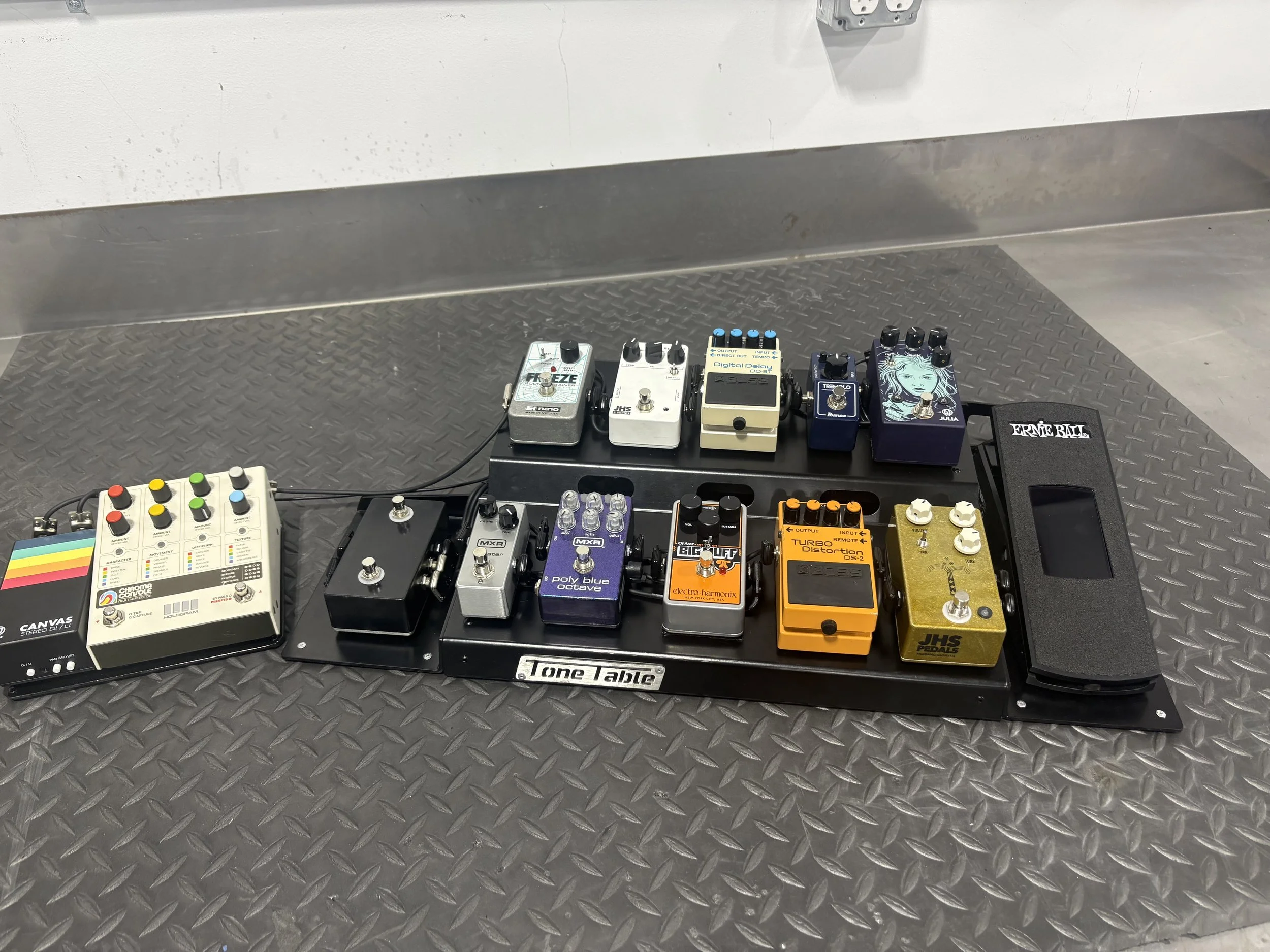Guitar effects pedalboard with multiple pedals including distortion, delay, reverb, and modulation effects on a black pedalboard labeled 'Tone Table' on a metal surface.