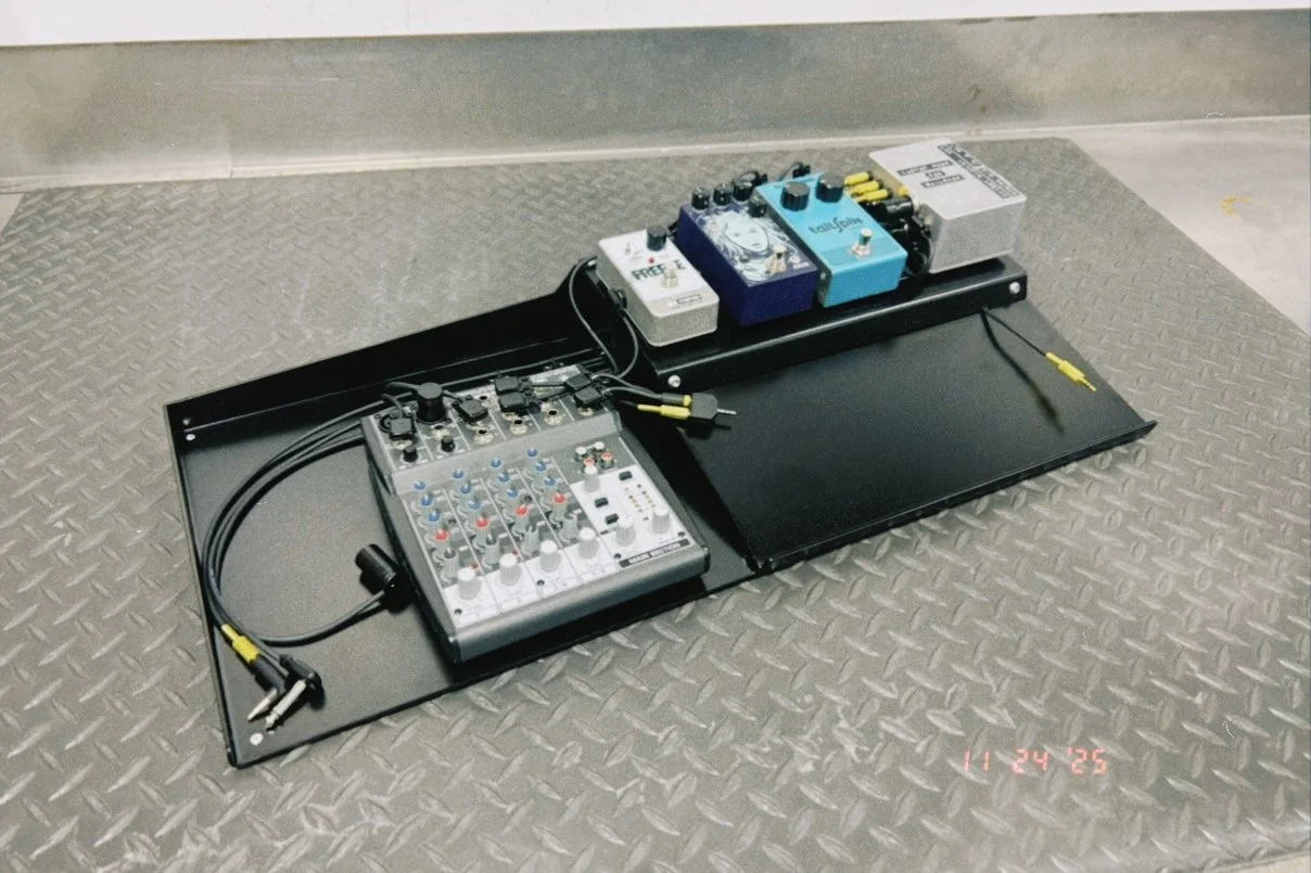A black pedalboard with multiple guitar effect pedals, an audio mixer, and various cables placed on a checkered metal floor.