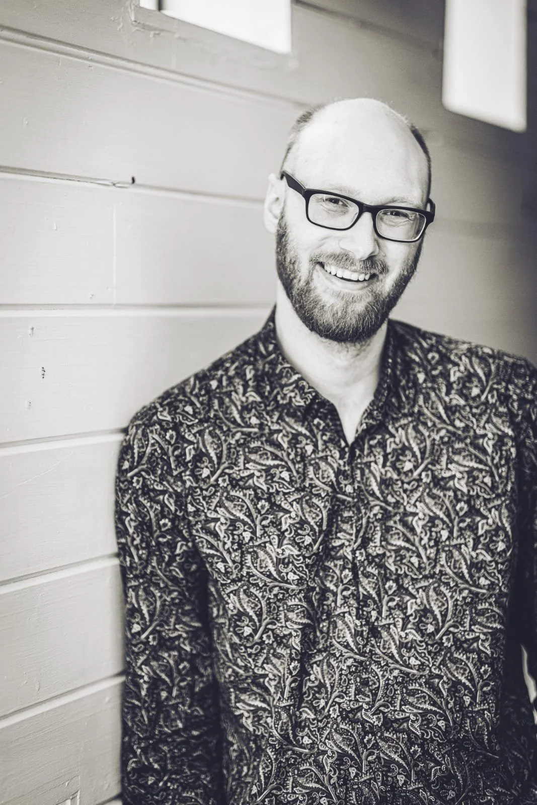 John, Community Management Lead at The Path, smiling with glasses and a beard, wearing a floral patterned shirt, leaning against a wooden wall.
