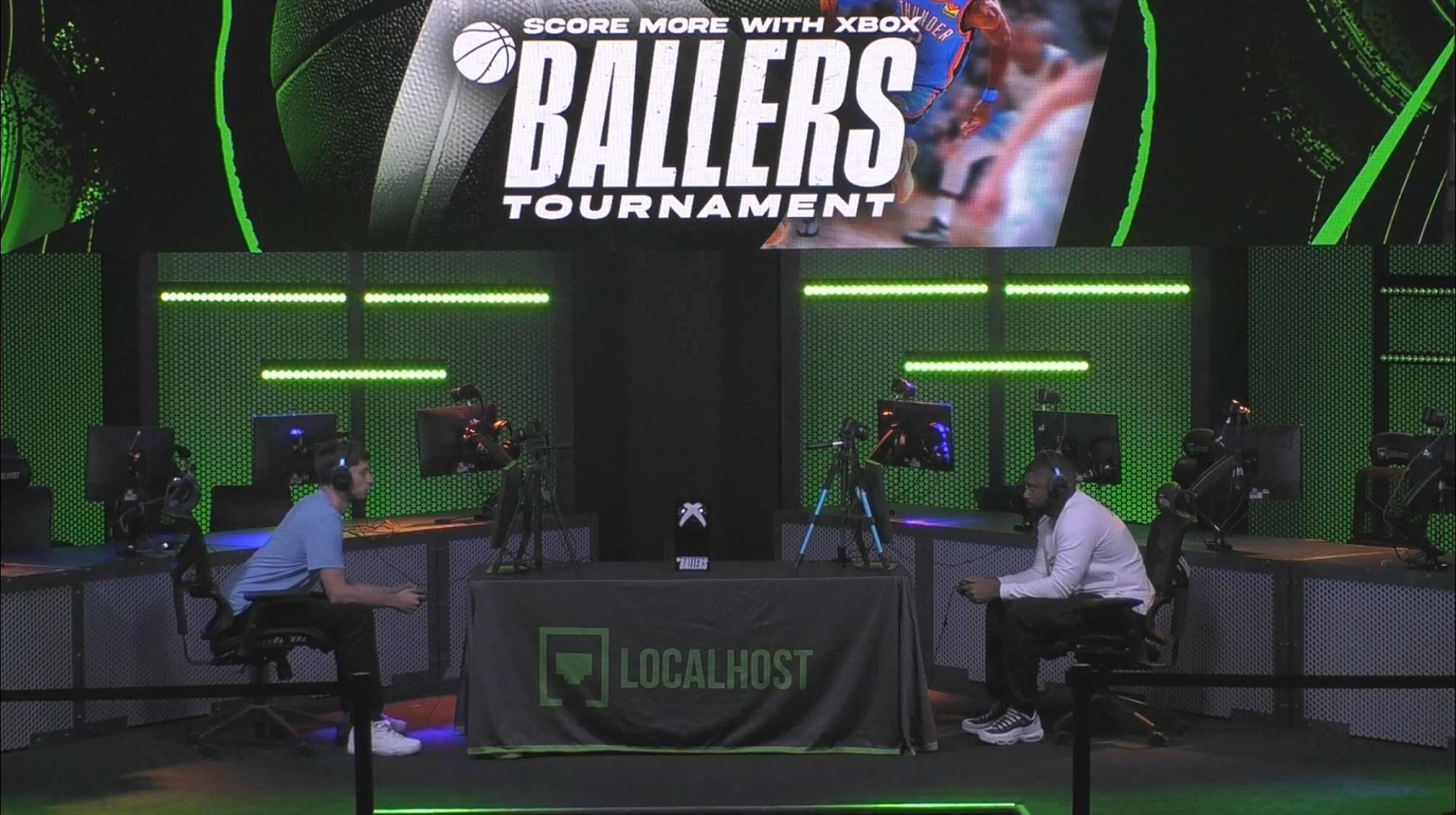 An esports tournament setup with two players sitting at tables facing each other, playing on desktop computers with gaming monitors, in a room with green and black lighting. A large screen above displays the event "Ballers Tournament" with basketball imagery.