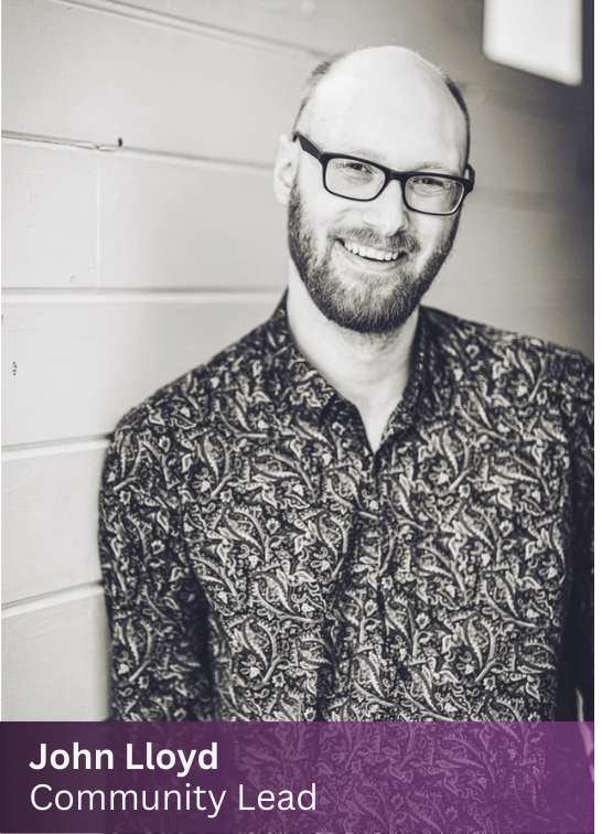 John, Community Management Lead at The Path, smiling with glasses and a beard, wearing a floral patterned shirt, leaning against a wooden wall.