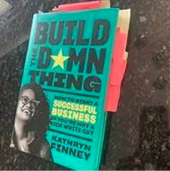 Stack of books with the top book titled 'Build the Damn Thing' by Kathryn Finney, featuring a photo of a woman wearing glasses on the cover.
