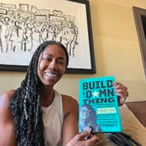 Woman with long braids holding a blue book titled 'Build Damn This' in a living room with a framed drawing on the wall.