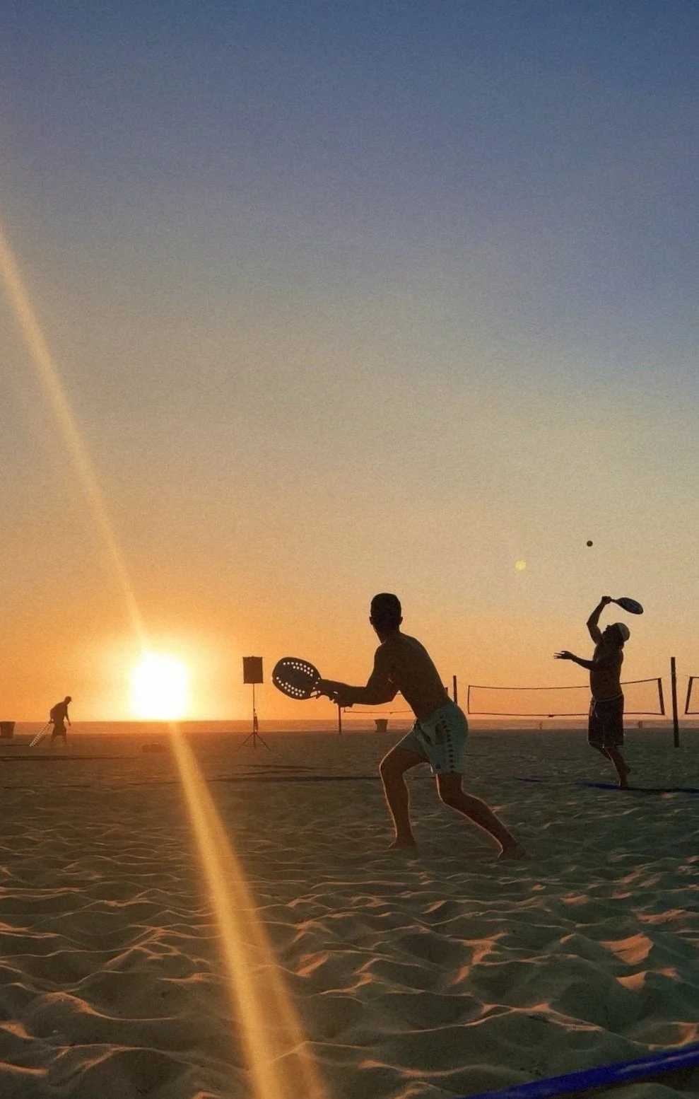 Silhouette of people playing beach tennis at sunset with a volleyball net in the background on a sandy beach.