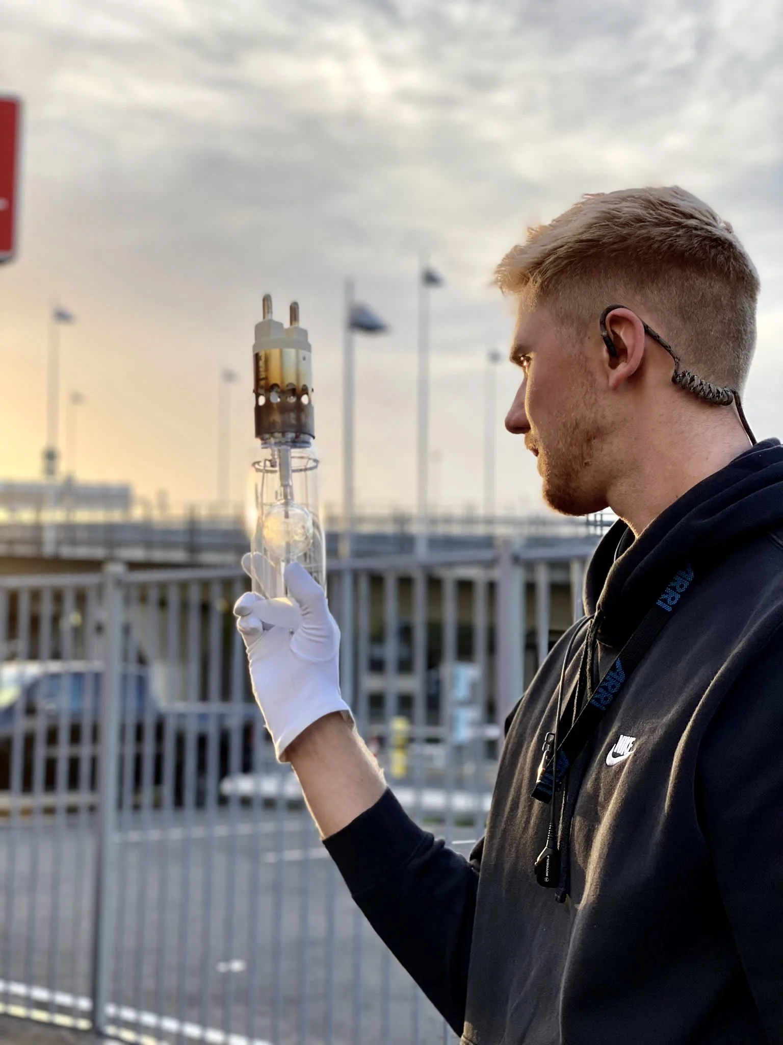 Ein Mann mit einem Ohrhörer hält eine Vorrichtung oder ein Gerät in der Hand, während er draußen steht. Er trägt einen schwarzen Pullover mit einem Logo und weiße Handschuhe. Der Hintergrund zeigt einen Zaun und unscharfe urbane Strukturen bei Sonnen