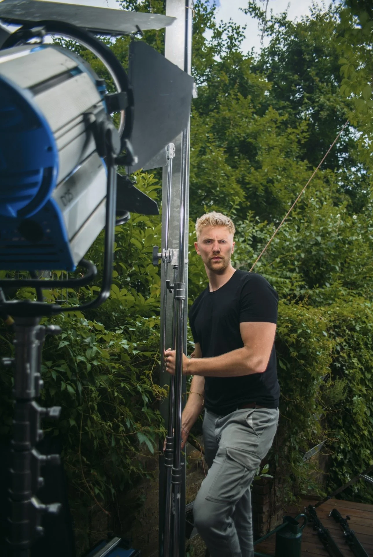 Ein Mann in einem schwarzen T-Shirt steht im Freien neben einer großen Filmleuchte vor einer mit Bäumen und Sträuchern bewachsenen Kulisse.