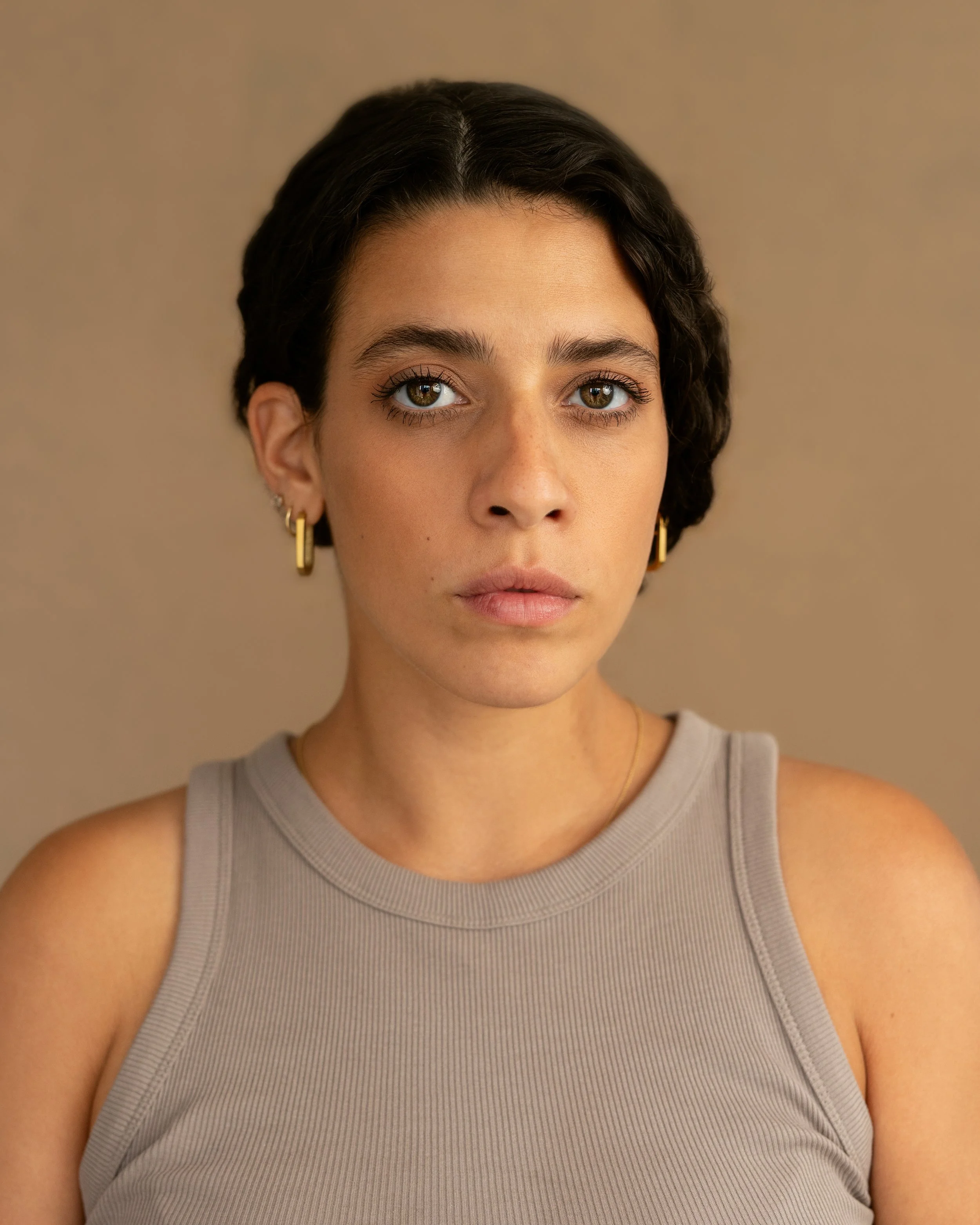 Close-up portrait of a young woman with short dark hair, wearing a gray sleeveless top and small gold hoop earrings, looking directly at the camera with a neutral expression.