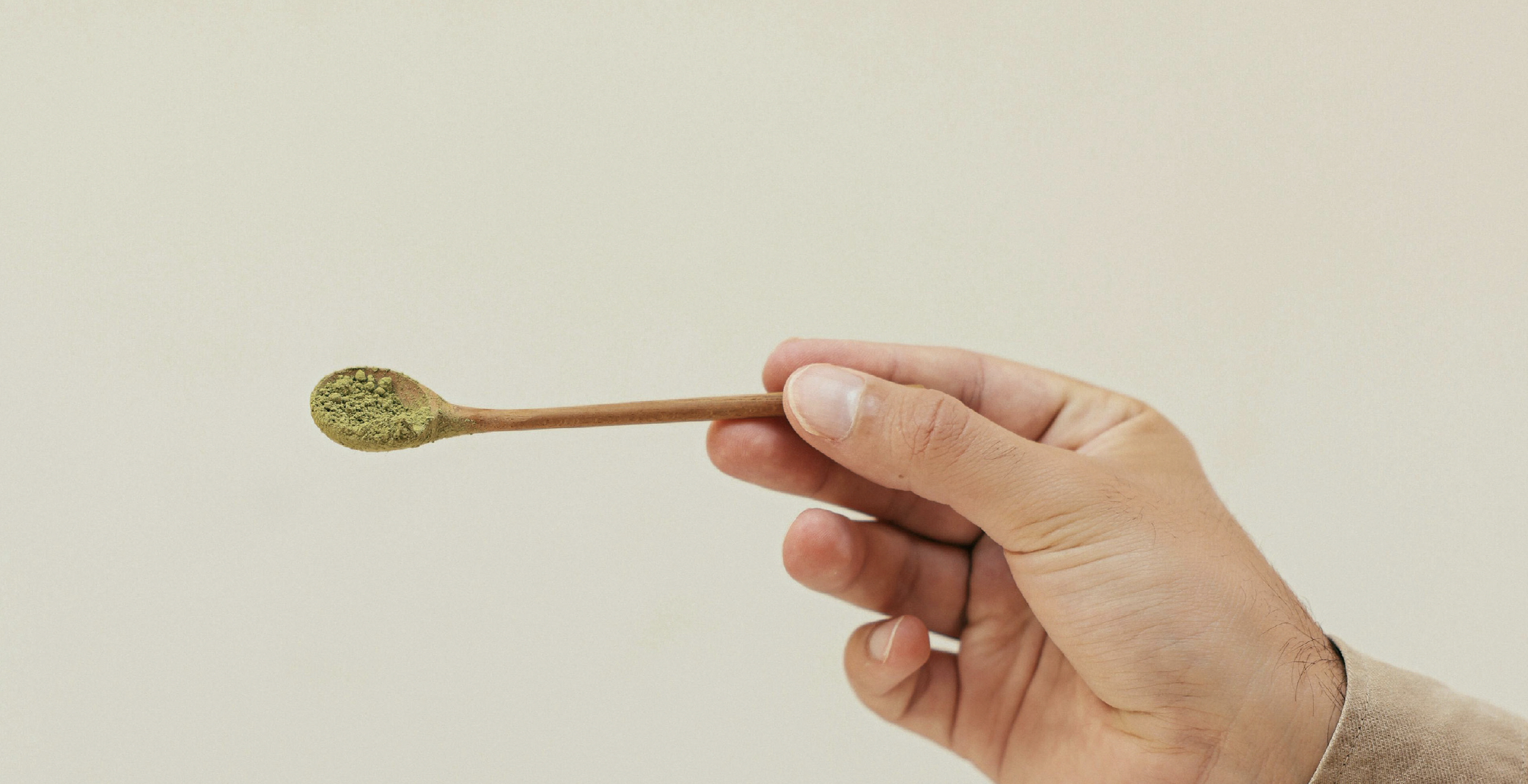 Hand holding a wooden stick with green powder on the end, against a plain beige background.