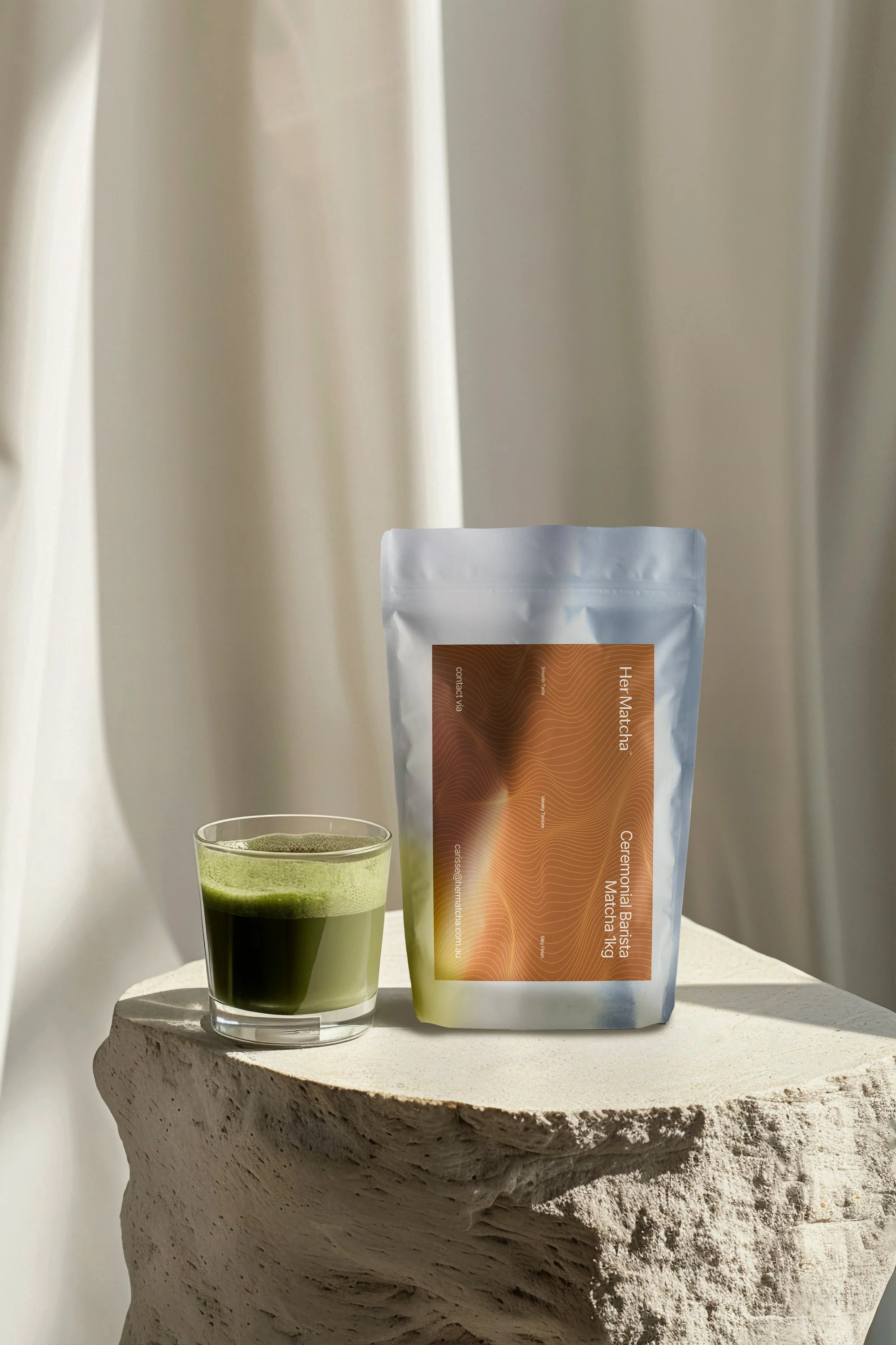 A glass of green matcha tea or powder next to a pouch of ceremonial matcha tea on a textured stone surface with a neutral background.