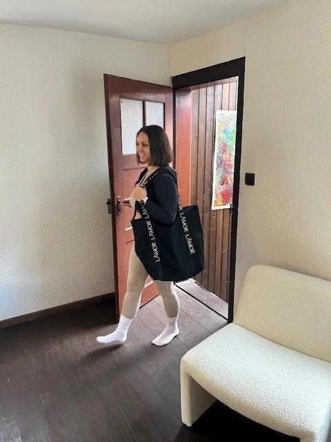 Frau mit dunklem Haar und dunkler Jacke, die eine große schwarze Tasche trägt, steht vor einer offenen Holzhaustür in einer Wohnung