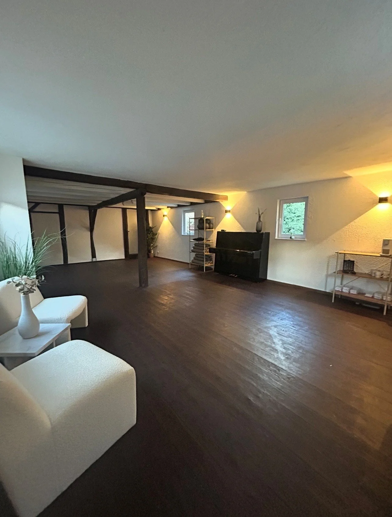 Geräumiges Wohnzimmer mit dunklem Holzboden, weißer Couch, schwarzen Klavier, Bücherregal, Pflanzen und gemütlicher Beleuchtung, mit kleinen Fenstern an der Wand.