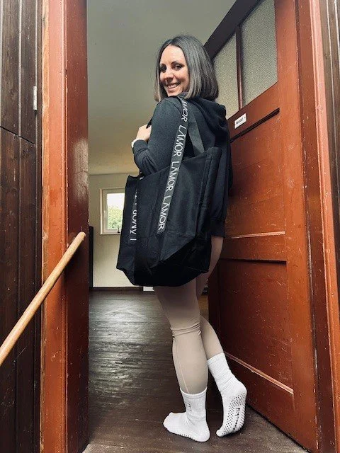 Eine lächelnde Frau steht im Türrahmen eines Hauses, trägt einen schwarzen Rucksack, ein dunkelblaues Sweatshirt, beige Leggings und weiße Socken.