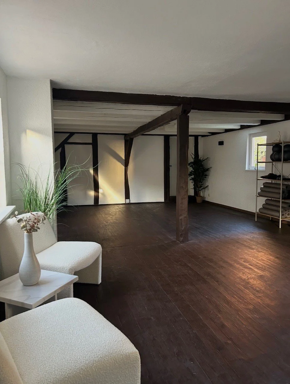 Ein ruhiger, heller Raum mit dunklem Holzfußboden, weißen Wänden, Holzstützen und einigen Pflanzen. Es gibt auch eine weiße Couch mit Beistelltisch und eine Regal mit Yogamatten und Fitnesszubehör.