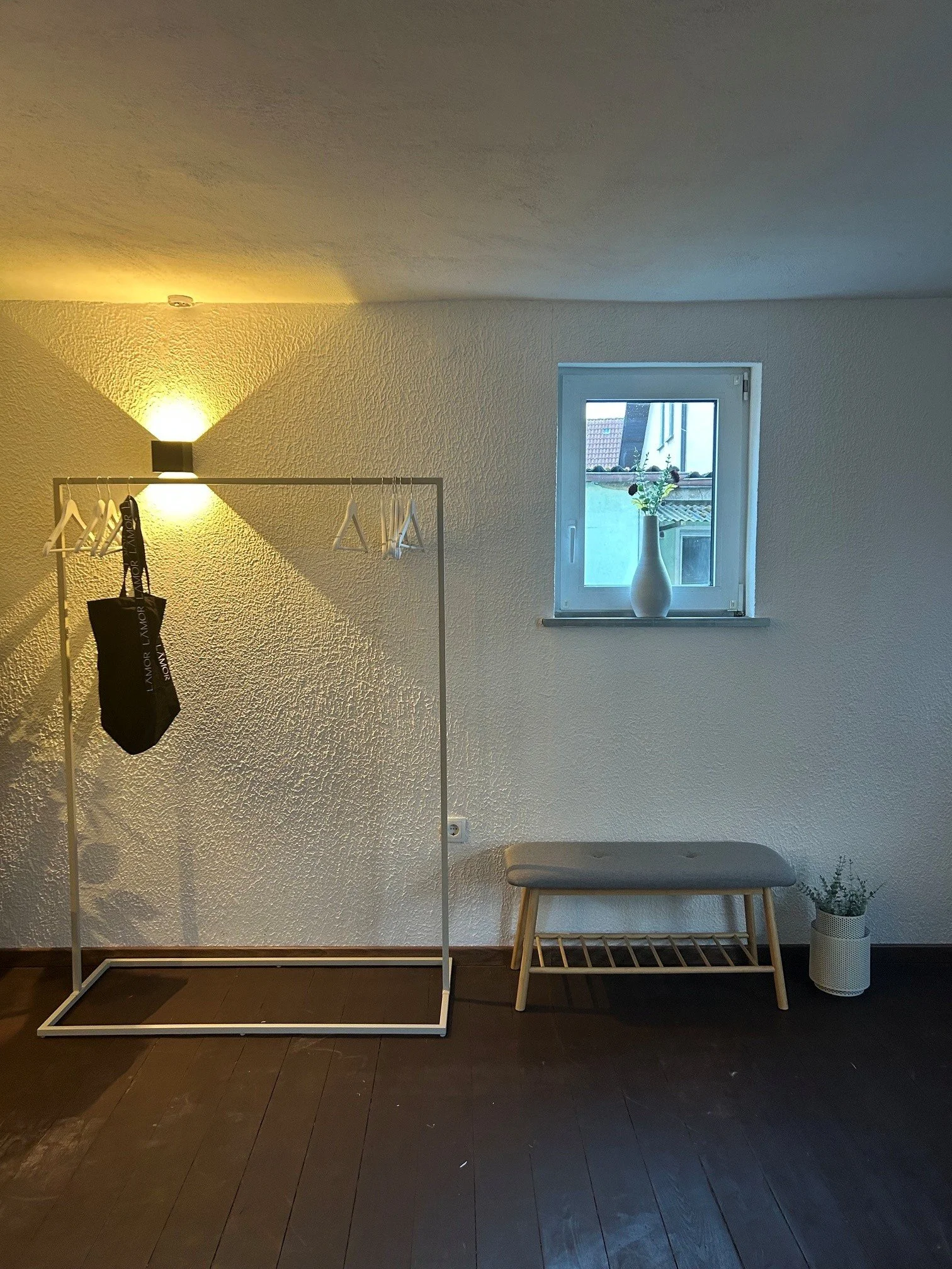 Innenraum mit weißem Wandschirm, Stehlampe, Fenster mit Vase und Blume, Hocker und Blumentopf mit Pflanze.