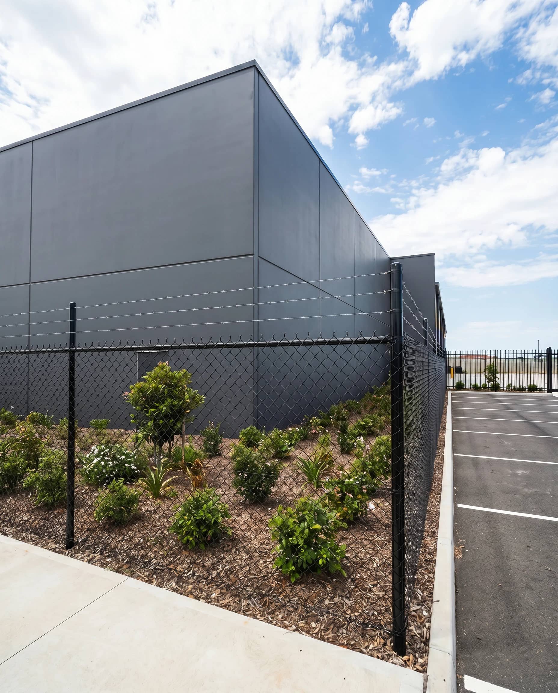 Corner angle of black chainwire fencing with barbed wire topping secured to commercial building exterior with landscaped garden bed