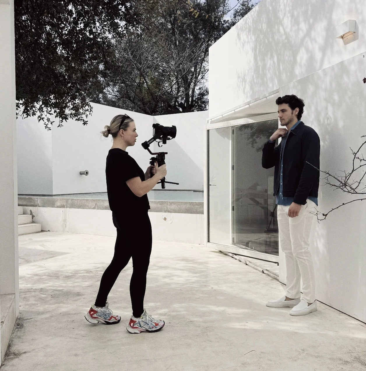 A woman filming a man with a camera stabilizer outside in a white-walled patio area.