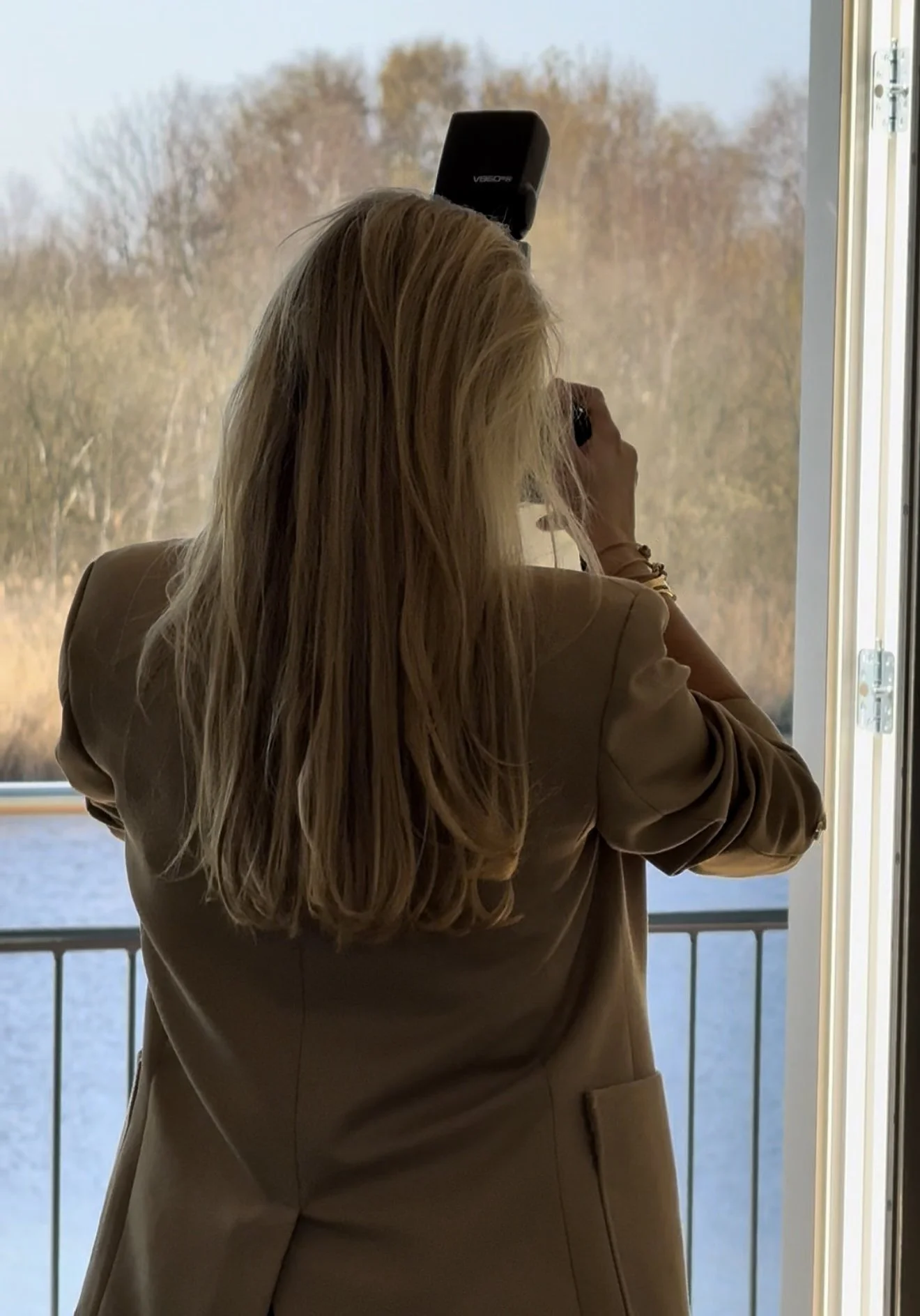 A woman with long blonde hair taking a photo with her phone near a window overlooking a body of water and trees.