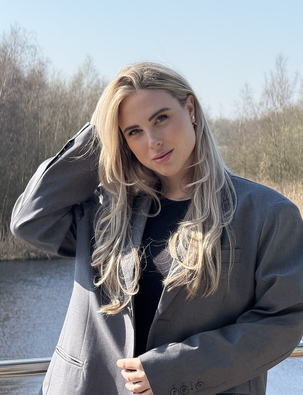 A young woman with long blonde hair, wearing a dark blazer, posing outdoors near a body of water with trees in the background on a sunny day.