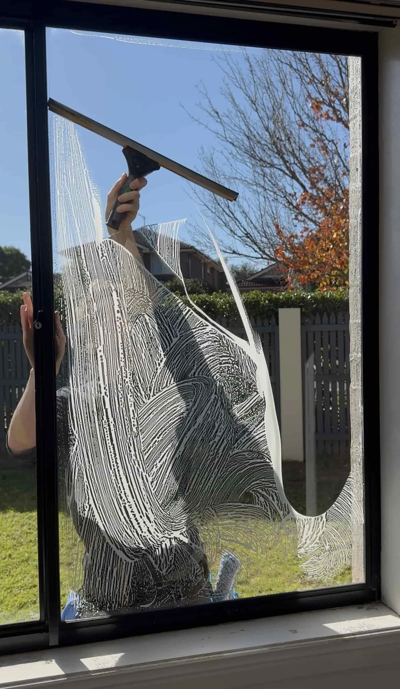 Person cleaning a window with a squeegee, leaving streaks and soap markings on glass, with houses and trees in the background.