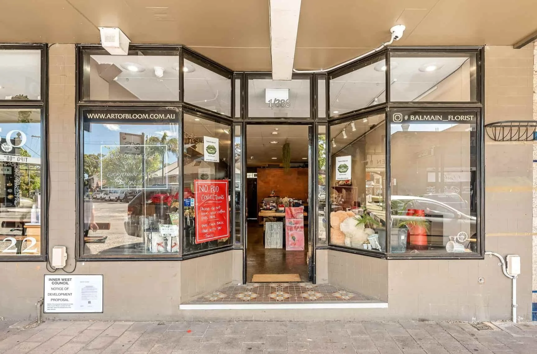 storefront of a florist shop with glass windows and door, displaying a website URL, social media handle, and a red sign that says 'No Pod Collections' inside the shop.