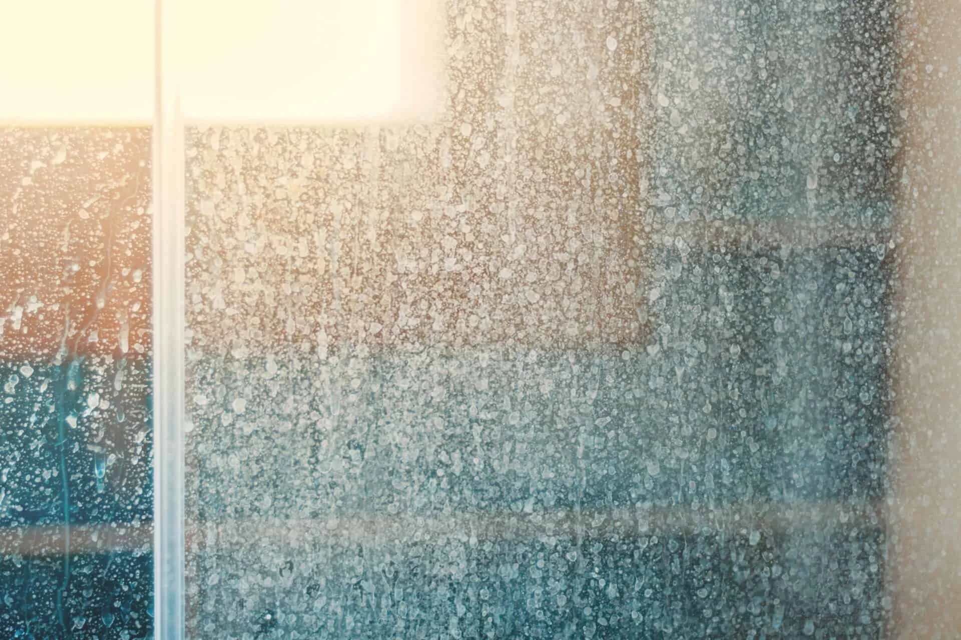 A foggy glass shower door with water droplets, partially revealing a bathroom interior with a lamp and tiles.