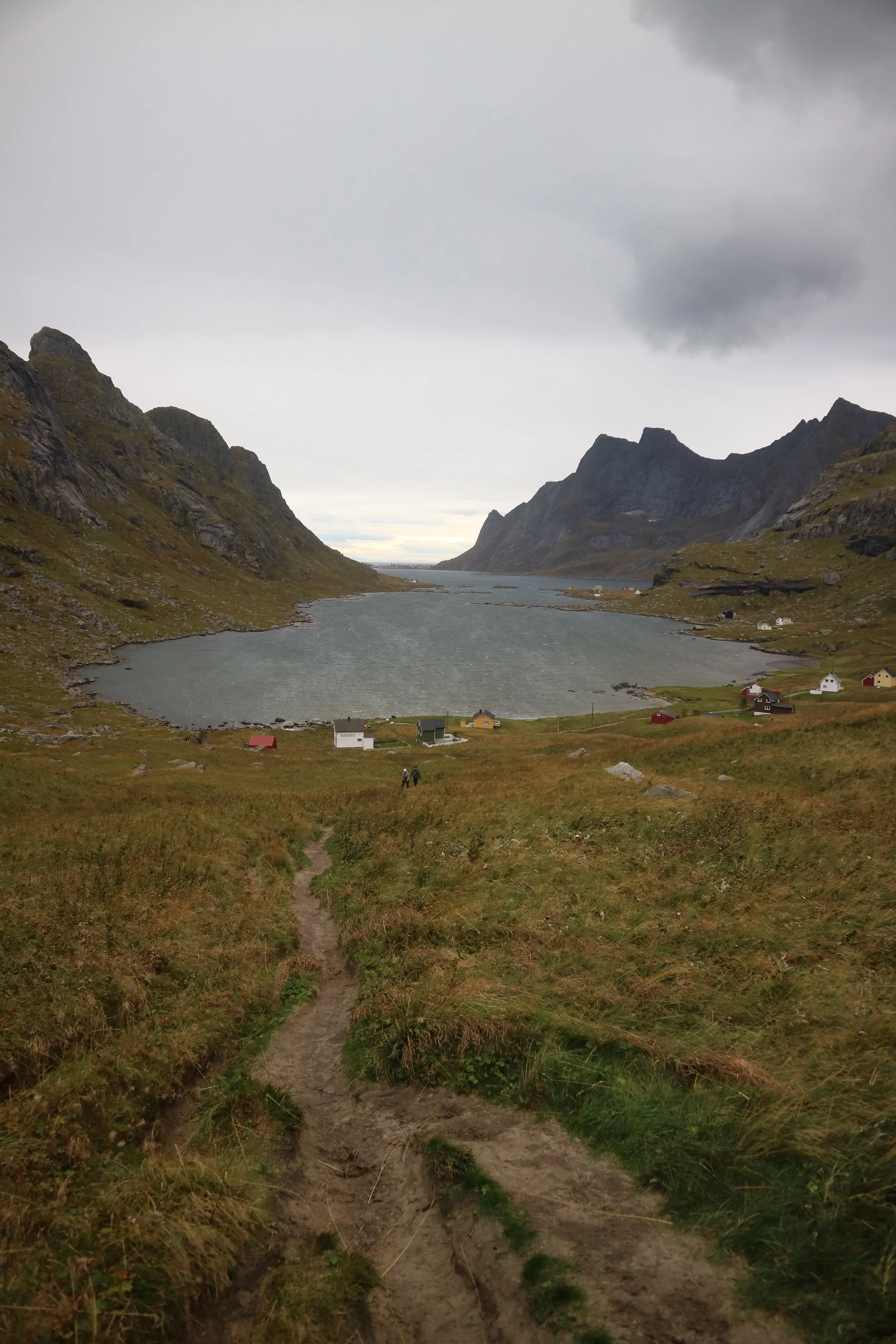 A scenic view of a thin, muddy, coastal like path to a Norwegian fjord surrounded by grassy hills and colourful houses on a cloudy day.