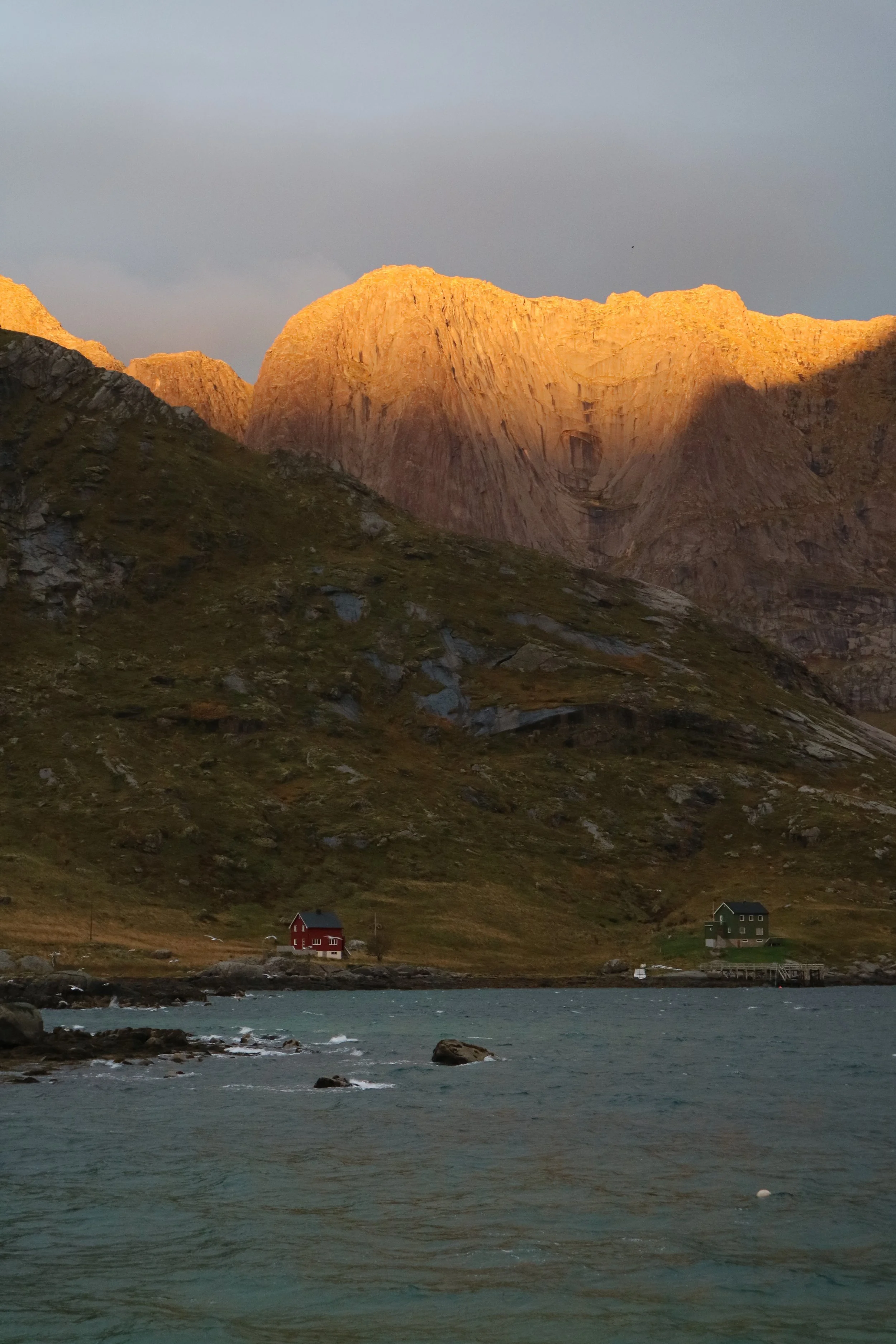 A scenic landscape with two houses, one green and one red, near fjord water at the base of large mountain cliffs illuminated by golden sunset.