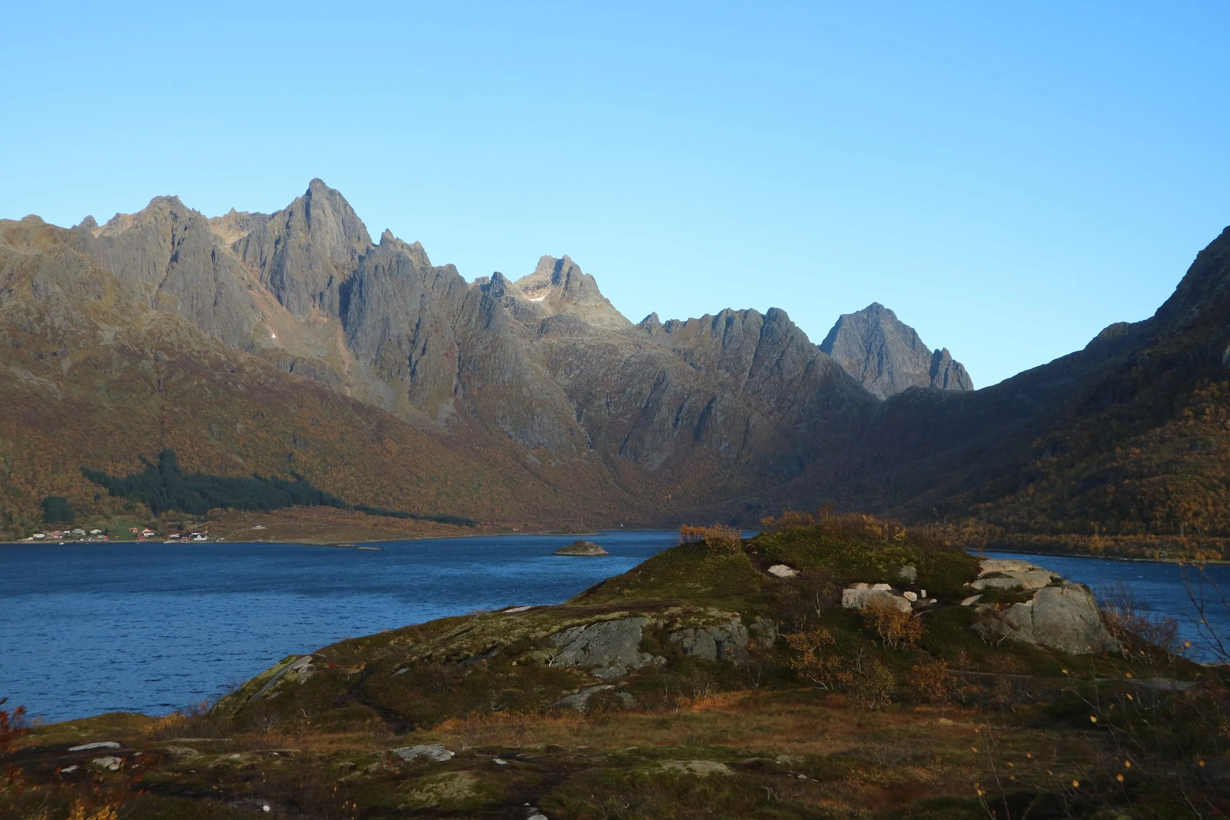 Lofoten, Norway, scenic landscape of a lake surrounded by mountains with rugged snow topped peaks, a small island, and autumnal coloured vegetation under a clear blue sky.