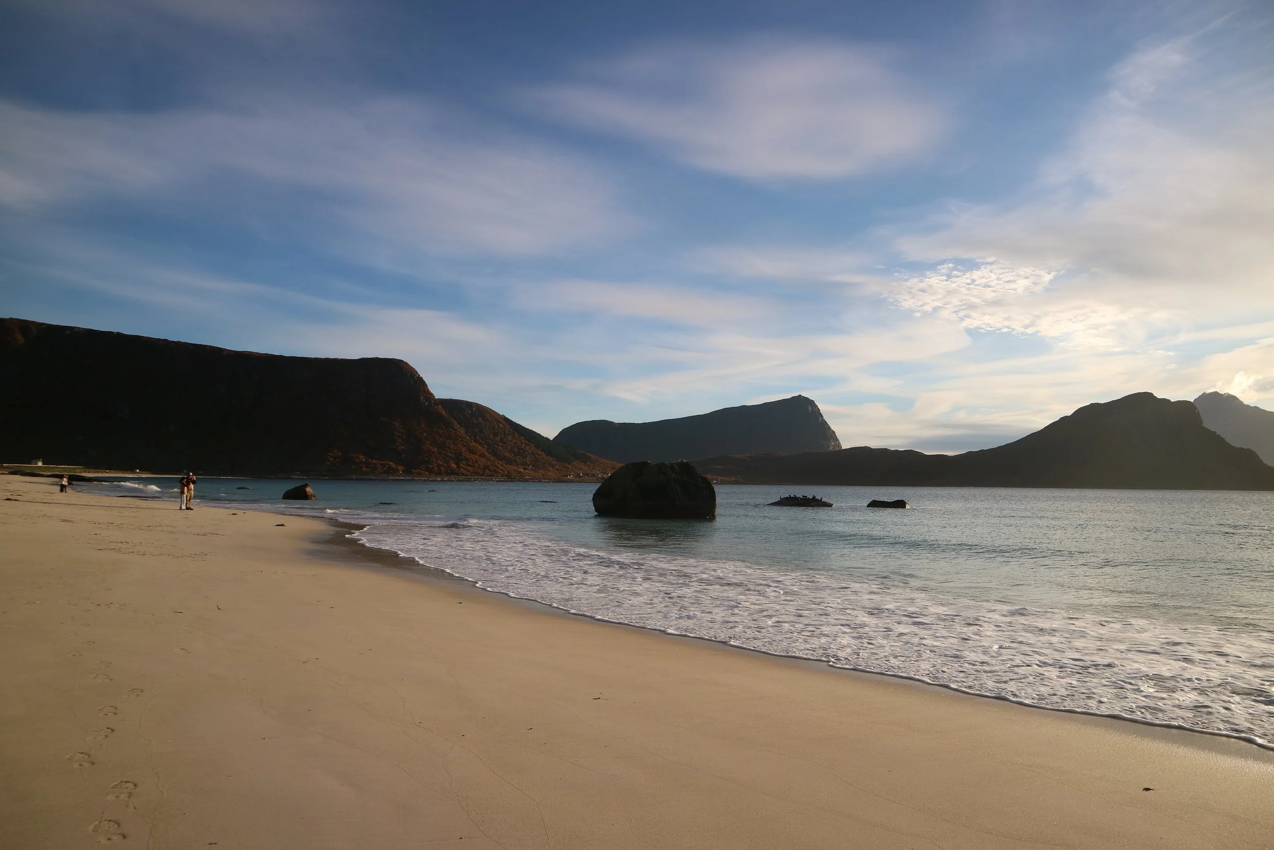 A sandy Haukland Beach, Norway, with large rocks in the water and mountains in the background under a blue sky with scattered clouds at golden hour.