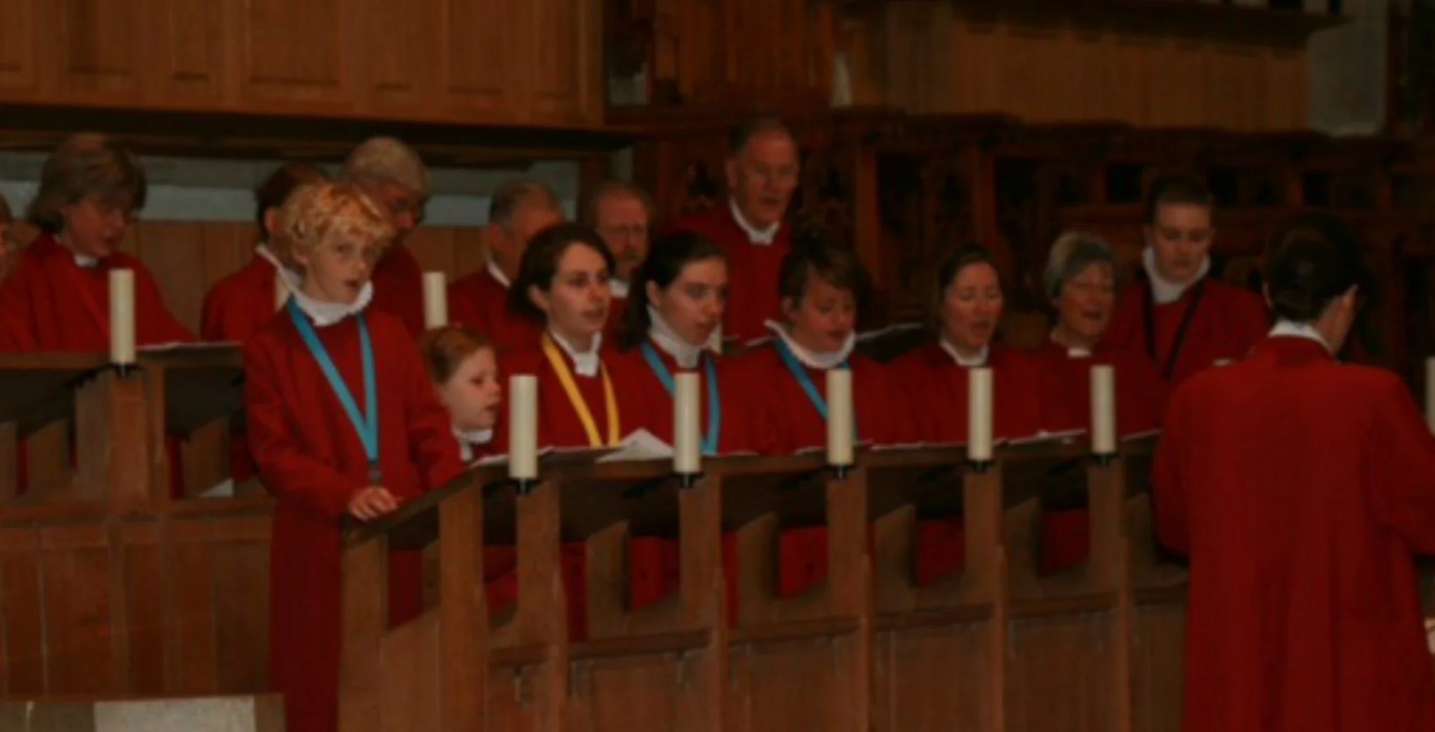 A choir dressed in burgundy cassock robes singing in a cathedral, all standing behind wooden choir stalls with white candles.