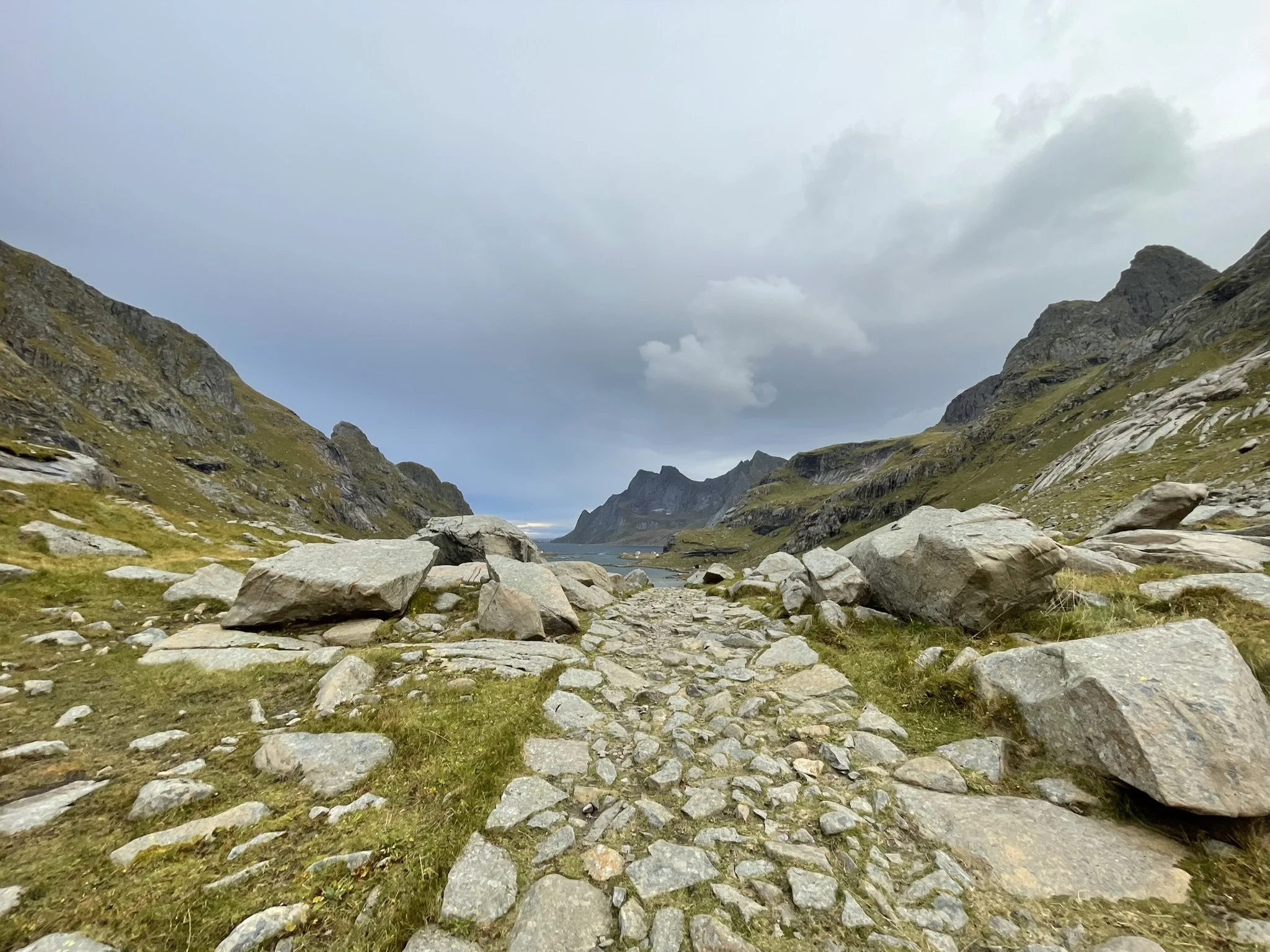 A rocky Norwegian mountain landscape with a cloudy sky, a narrow stone path with large rocks either side leading towards distant mountains and a lake.