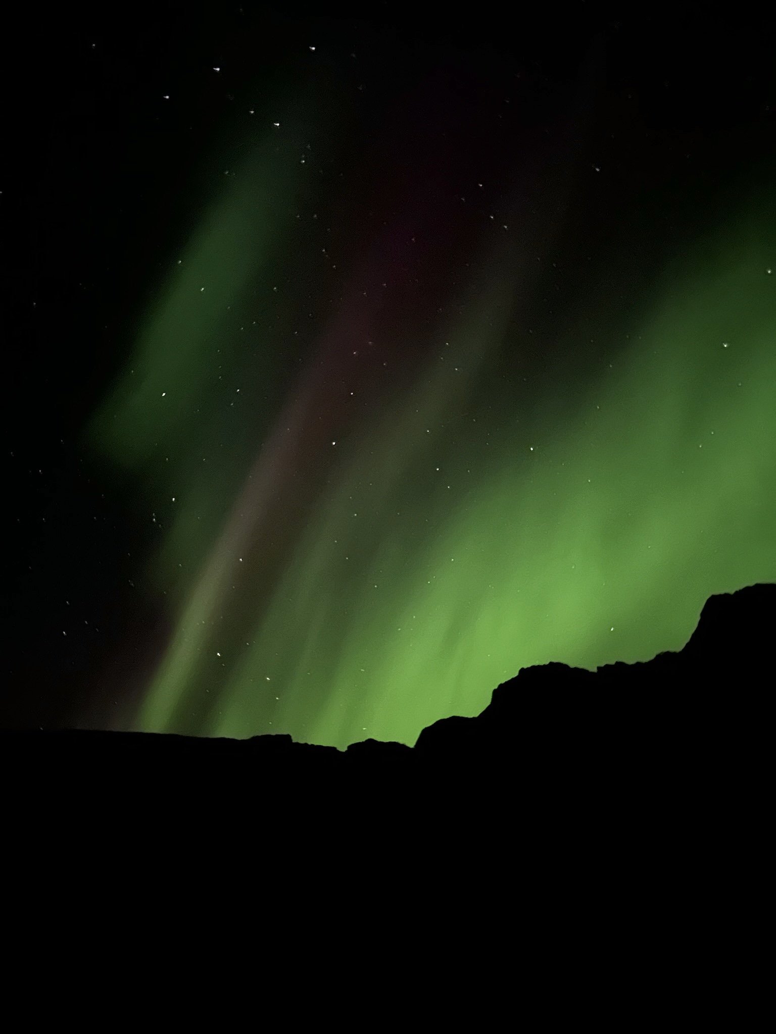 Northern lights, Aurora Borealis, in green, purple, and dusty maroon colours in the starry night sky over a black silhouette of a Norwegian mountain.