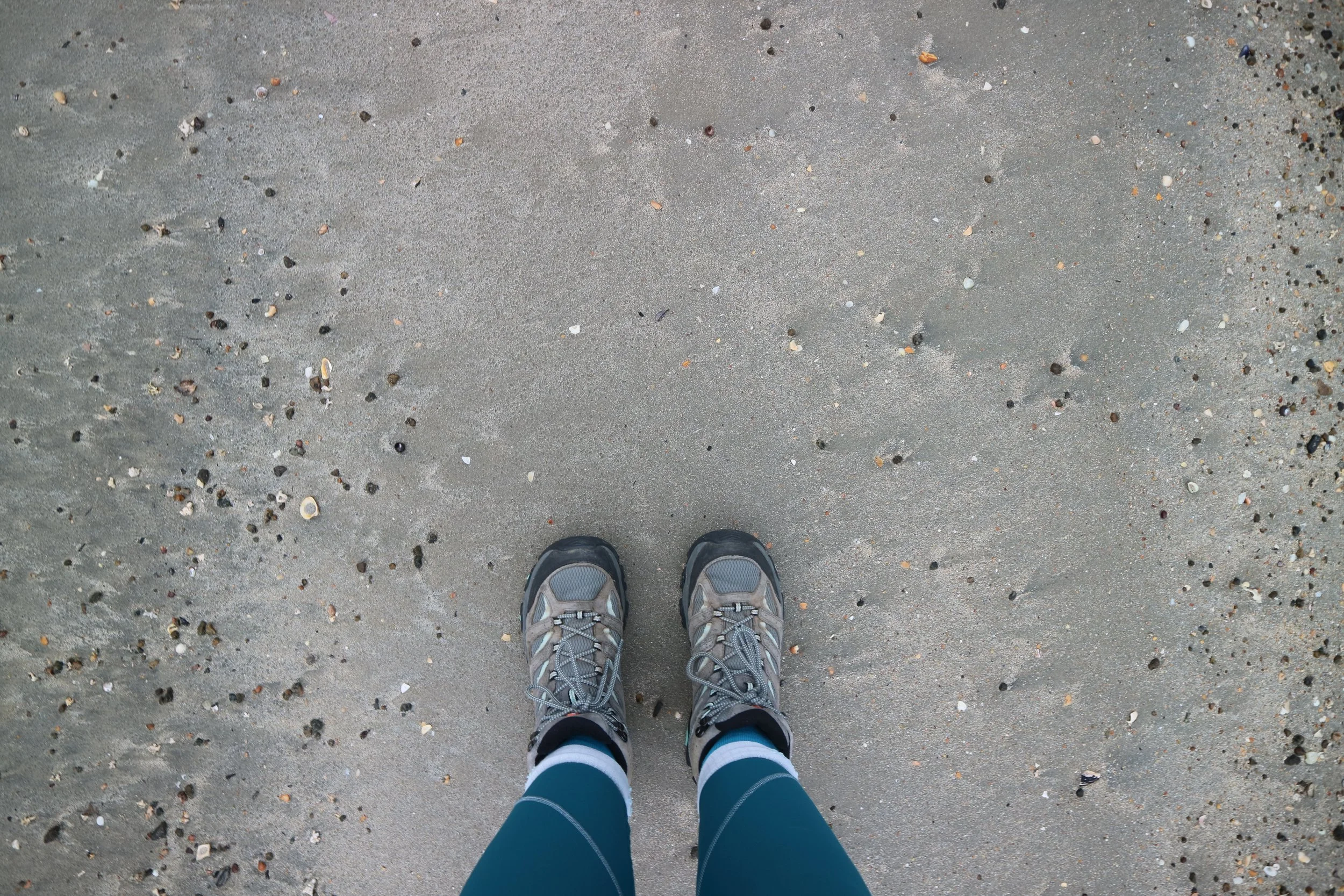 View from above of a person standing on a sandy beach, wearing grey and teal walking boots, turquoise leggings and socks, with small shells and pebbles scattered around.