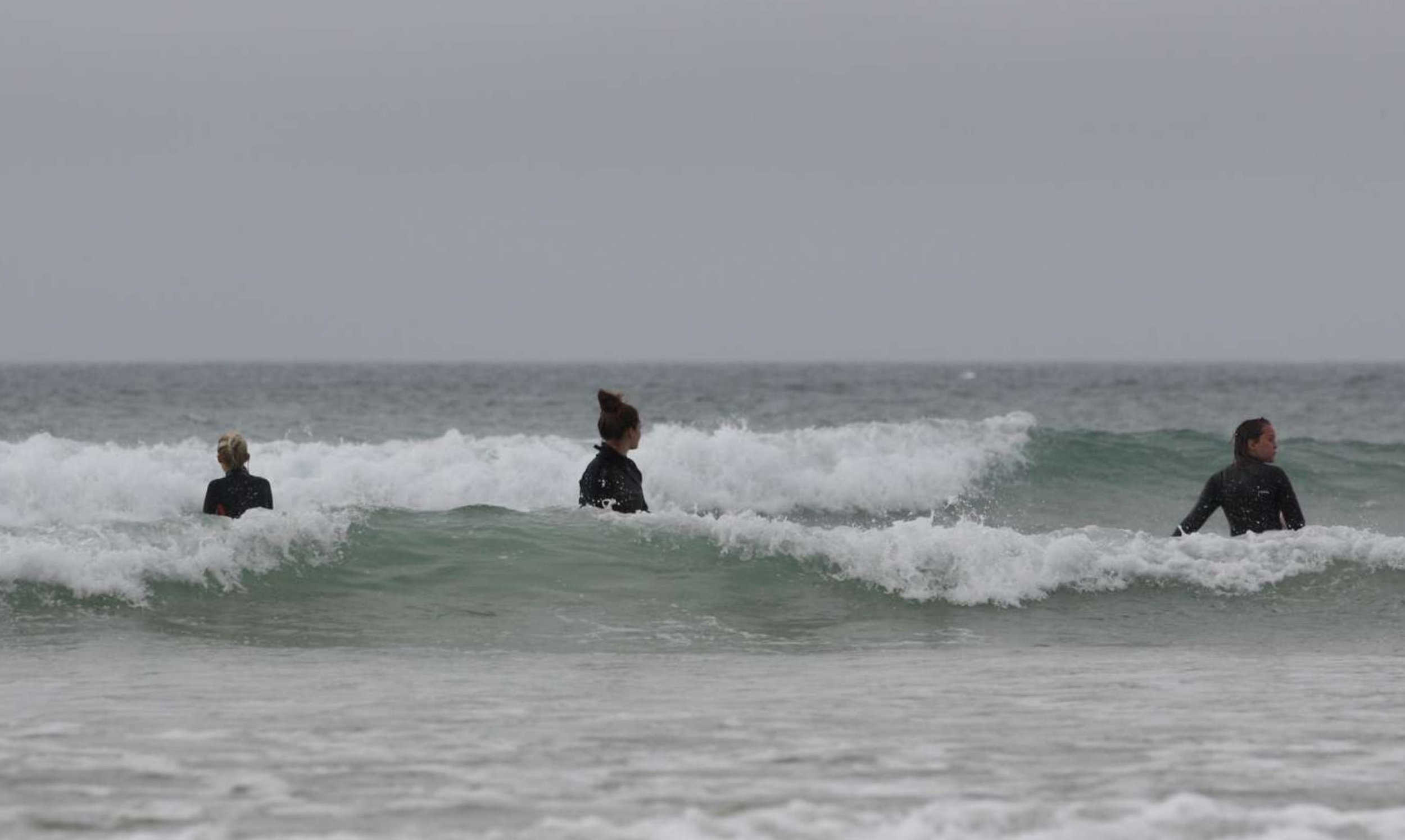 Three women are surfing in the ocean waves, where the photo is taken near the shoreline, each wearing black wetsuits, facing the water on an overcast day.
