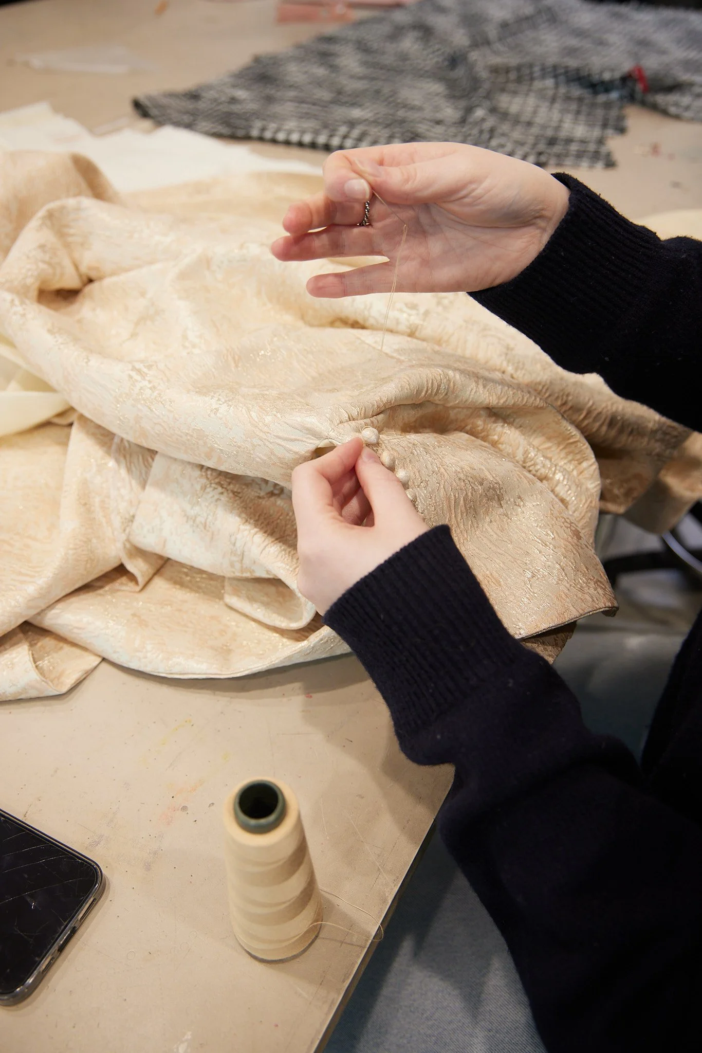 Person's hands sewing small rouleau buttons onto a champagne and blush, textured fabric using a needle and thread in a workspace.