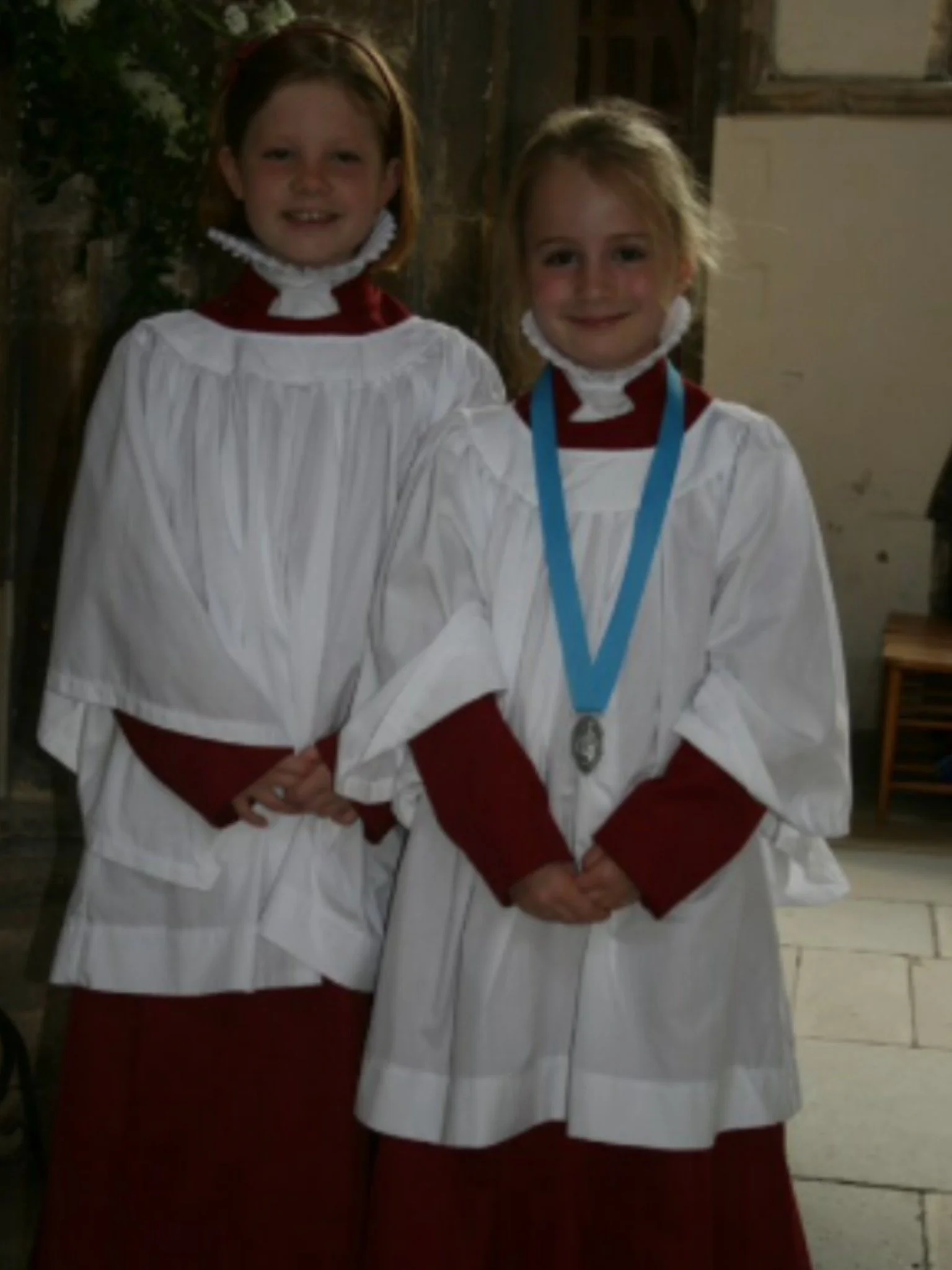 Two young girls dressed in white ruffs and surplices over burgundy cassocks, standing in the indoors of a cathedral. The younger girl has a blue medal around her neck. Both are smiling.
