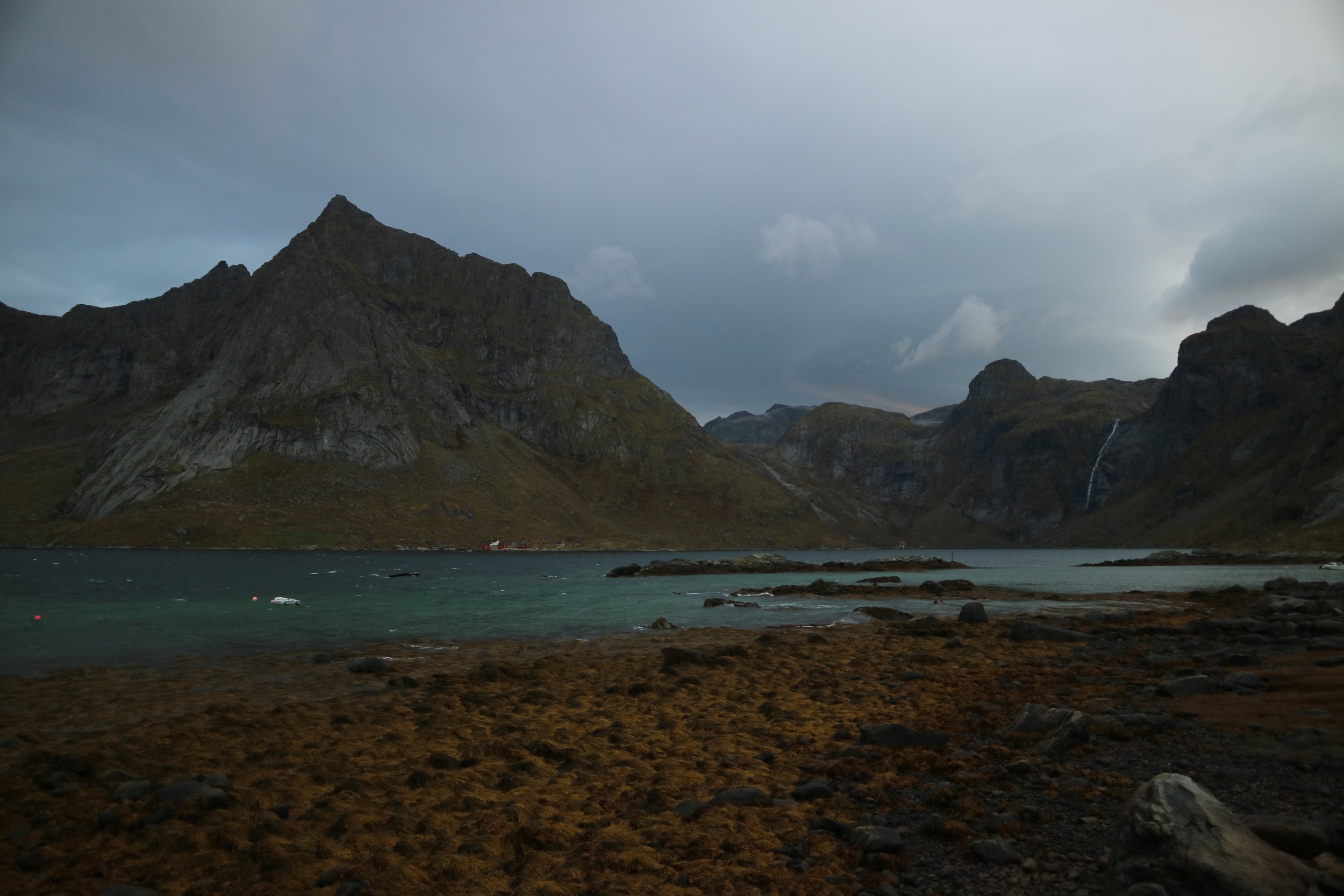 A Norwegian mountainous landscape with a lake in the foreground, rugged rocky terrain covered in seaweed at the waters edge, and overcast skies.