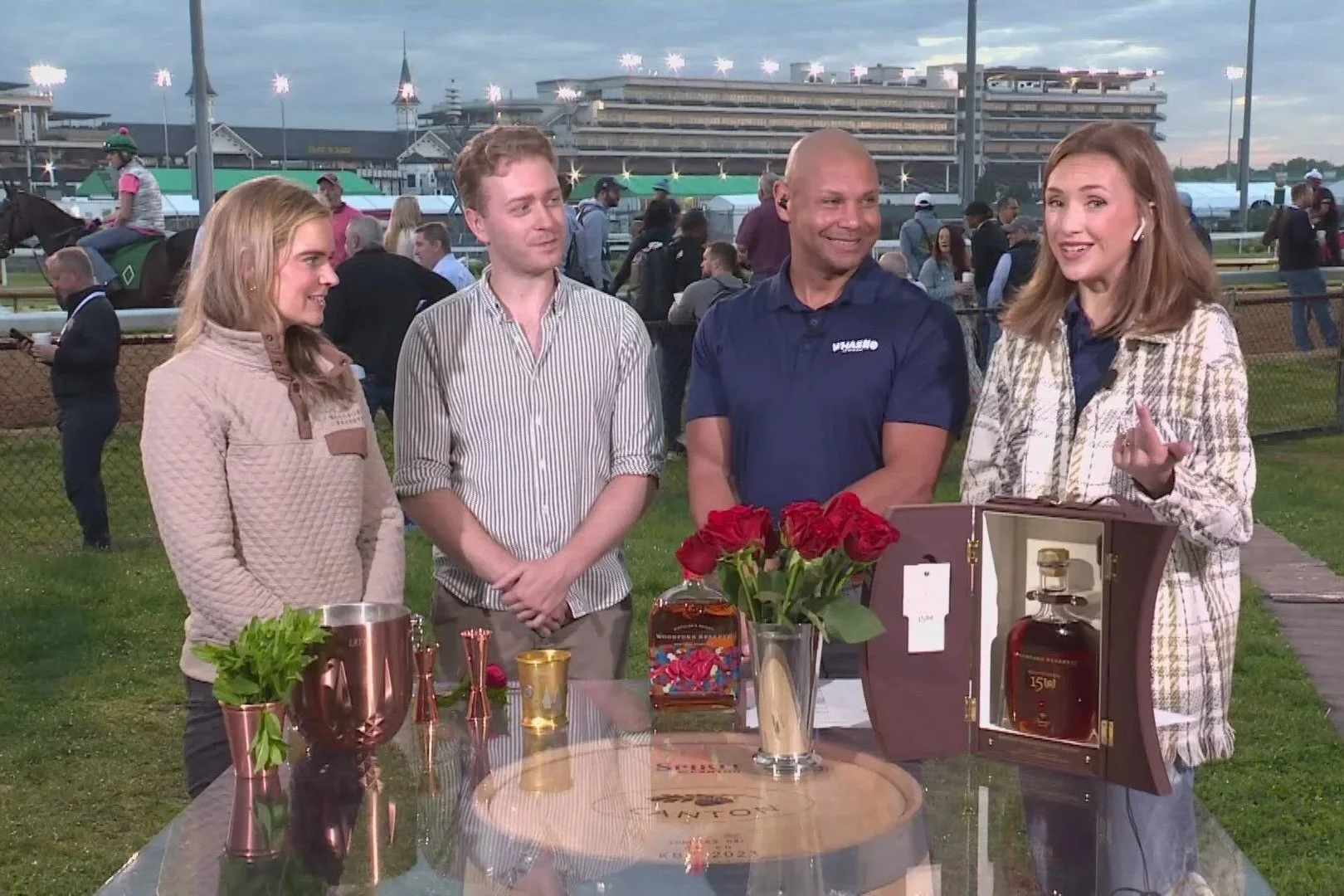 Group of four people standing around a table at a horse racing event, with a racecourse and spectators in the background. One woman is speaking and gesturing, with a whiskey bottle on the table.