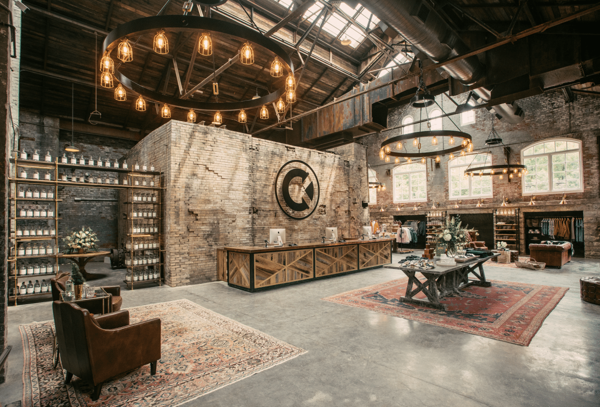 Interior of a stylish retail store with exposed brick walls, large windows, and industrial decor. There are circular chandeliers with Edison bulbs, a central wooden counter, clothing racks, and shelves displaying products. Seating areas with armchairs and rugs are also visible.