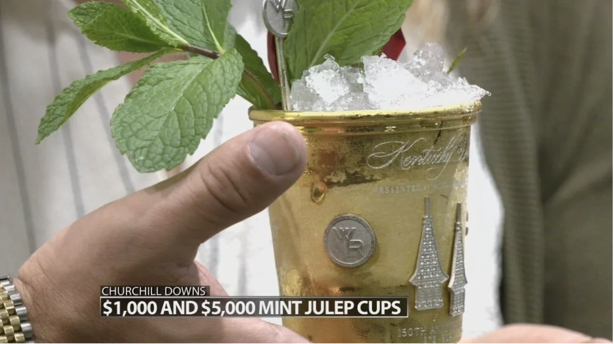 Close-up of a gold-colored cup with decorative elements, filled with a mint julep cocktail topped with crushed ice and mint leaves, with a hand holding it. The background shows part of a person's shirt and arm, with a subtitle at the bottom indicating the cup's value of $1,000 and $5,000 mint julep cups.