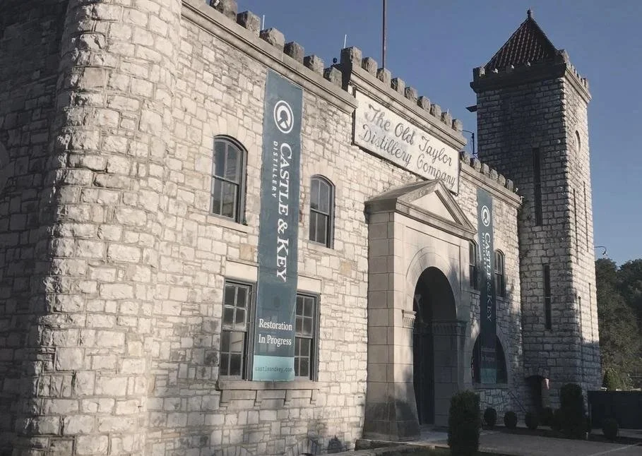 Exterior view of a historic stone castle with a tower, featuring banners for 'Castle & Key Distillery' and sign indicating 'Restoration in Progress.' The castle is labeled as 'The Old Taylor Distillery Company.'